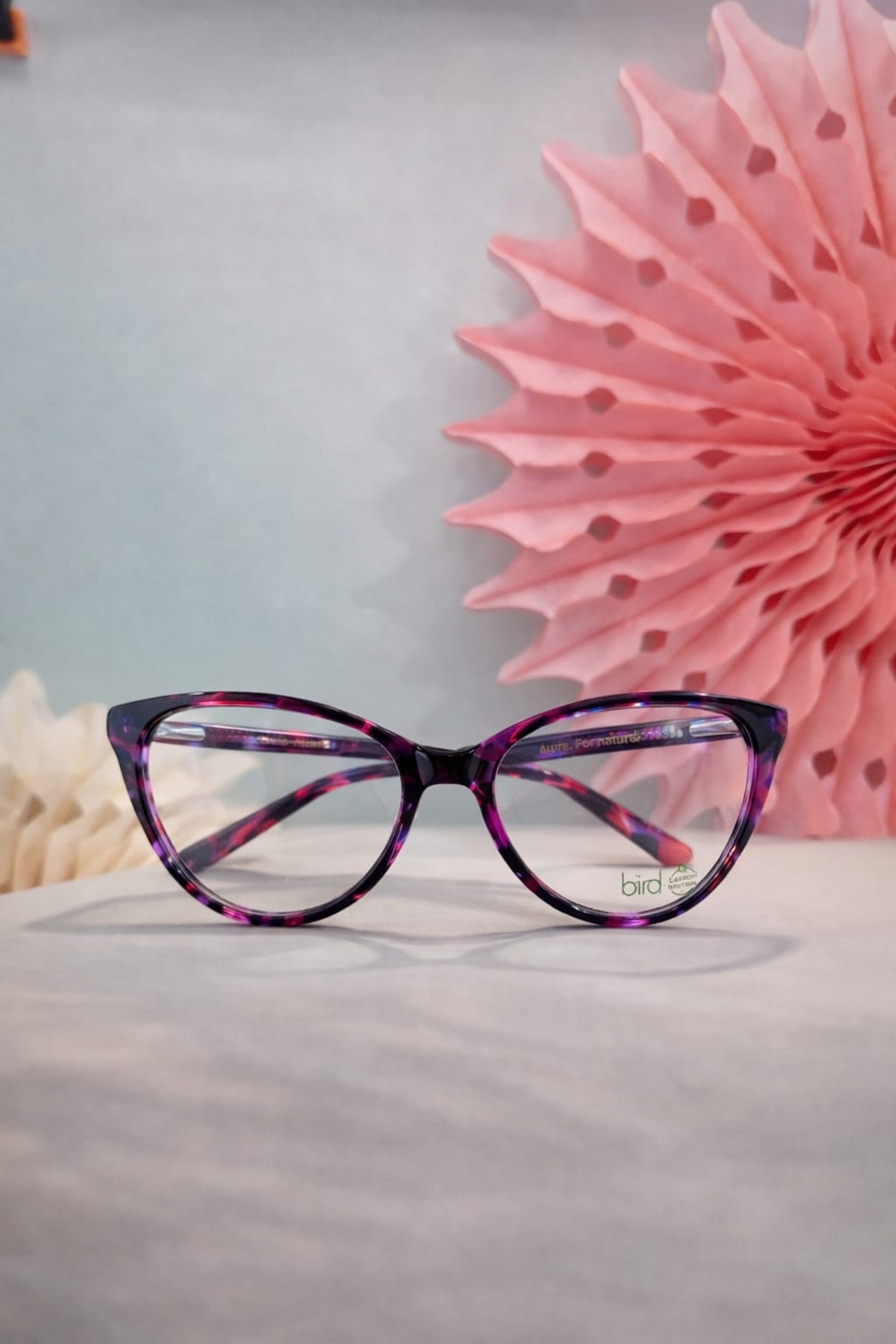 Purple patterned cat-eye eyeglass frames displayed against a soft pink decorative backdrop