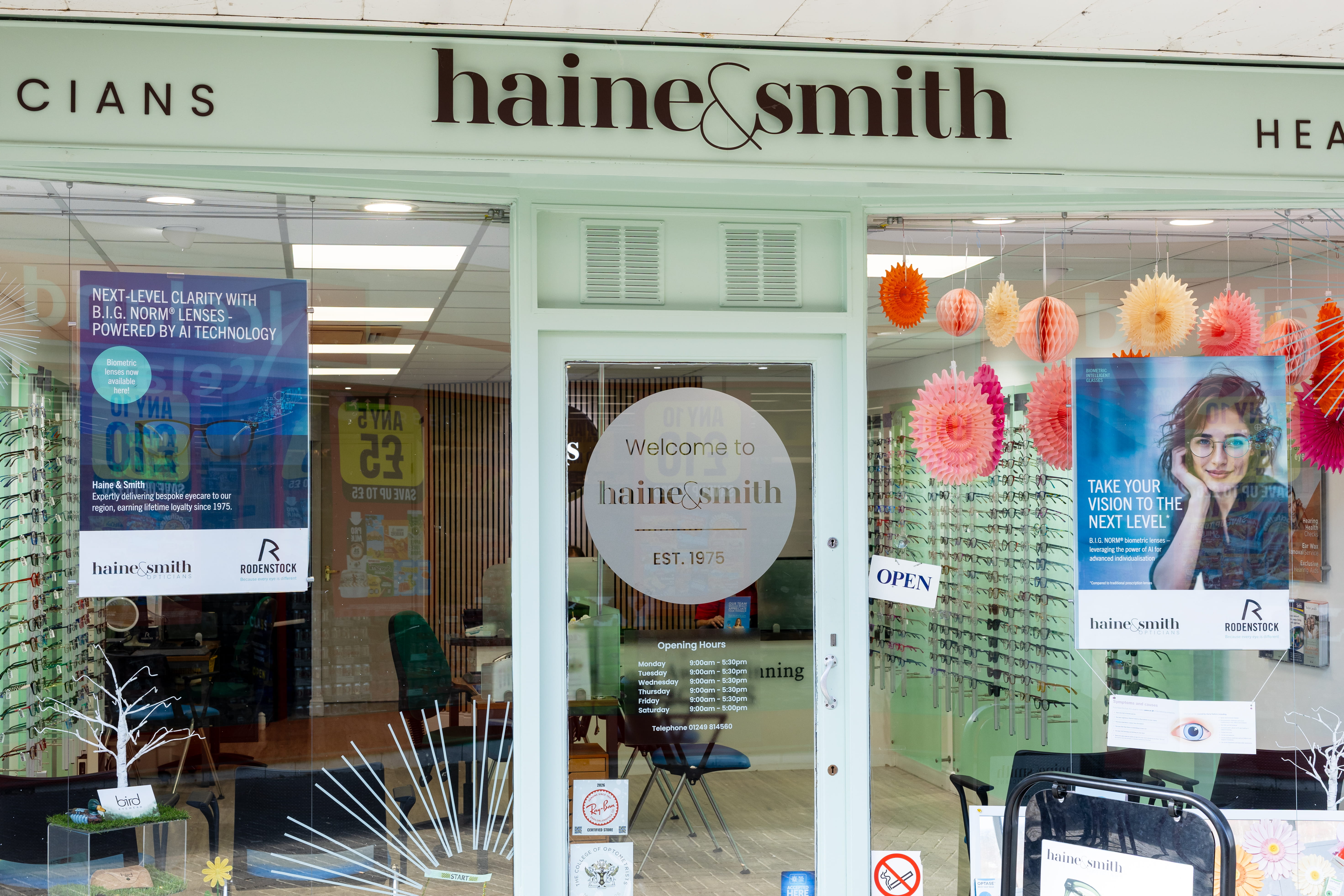 Front entrance of a Haine & Smith Opticians practice featuring large display windows with eyewear collections, promotional vision care posters, colourful window decorations, and a welcoming glass door displaying opening hours