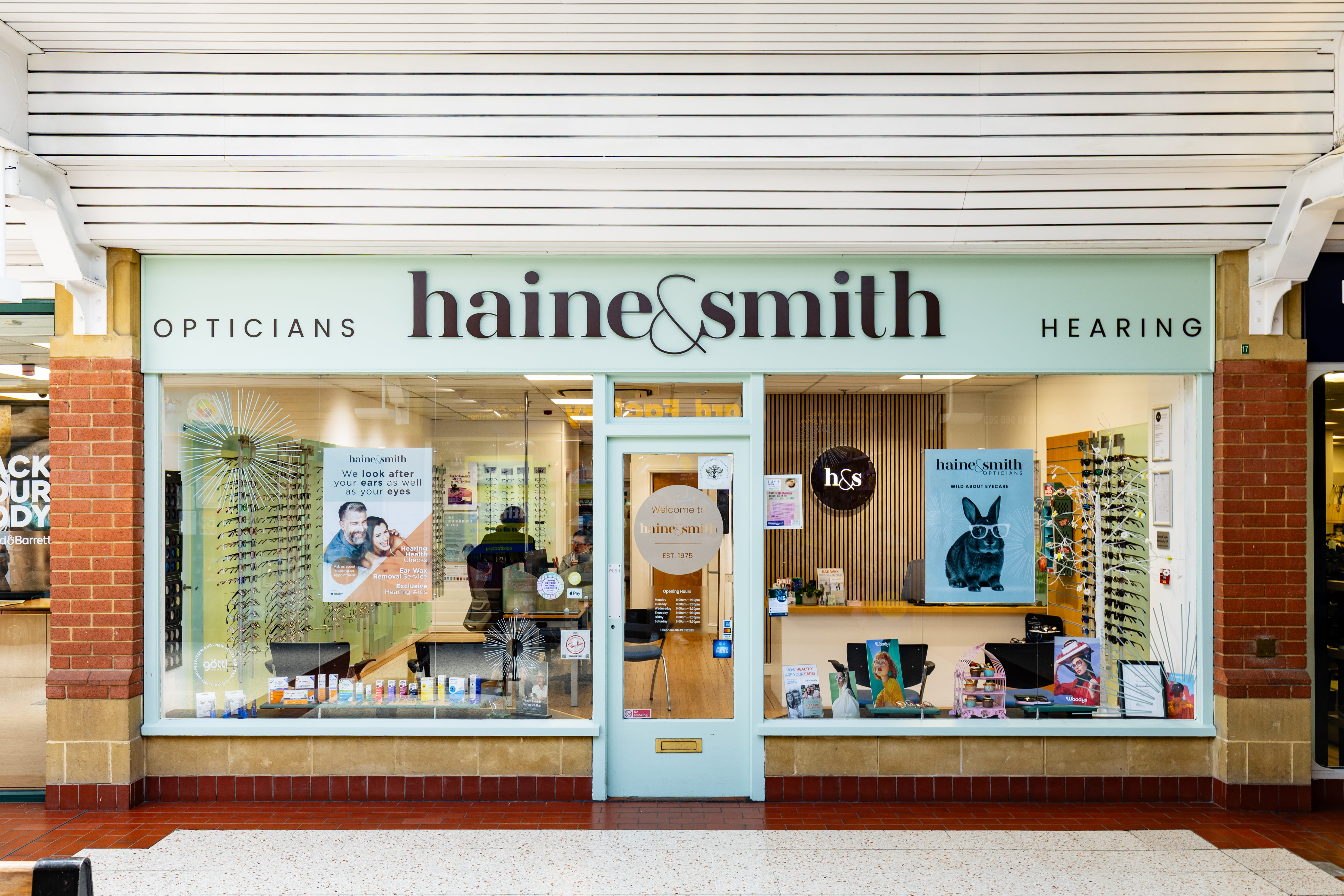 Exterior view of a Haine & Smith Opticians and Hearing Care practice in a shopping centre, featuring branded storefront signage, eyewear displays, and eye care products in the window