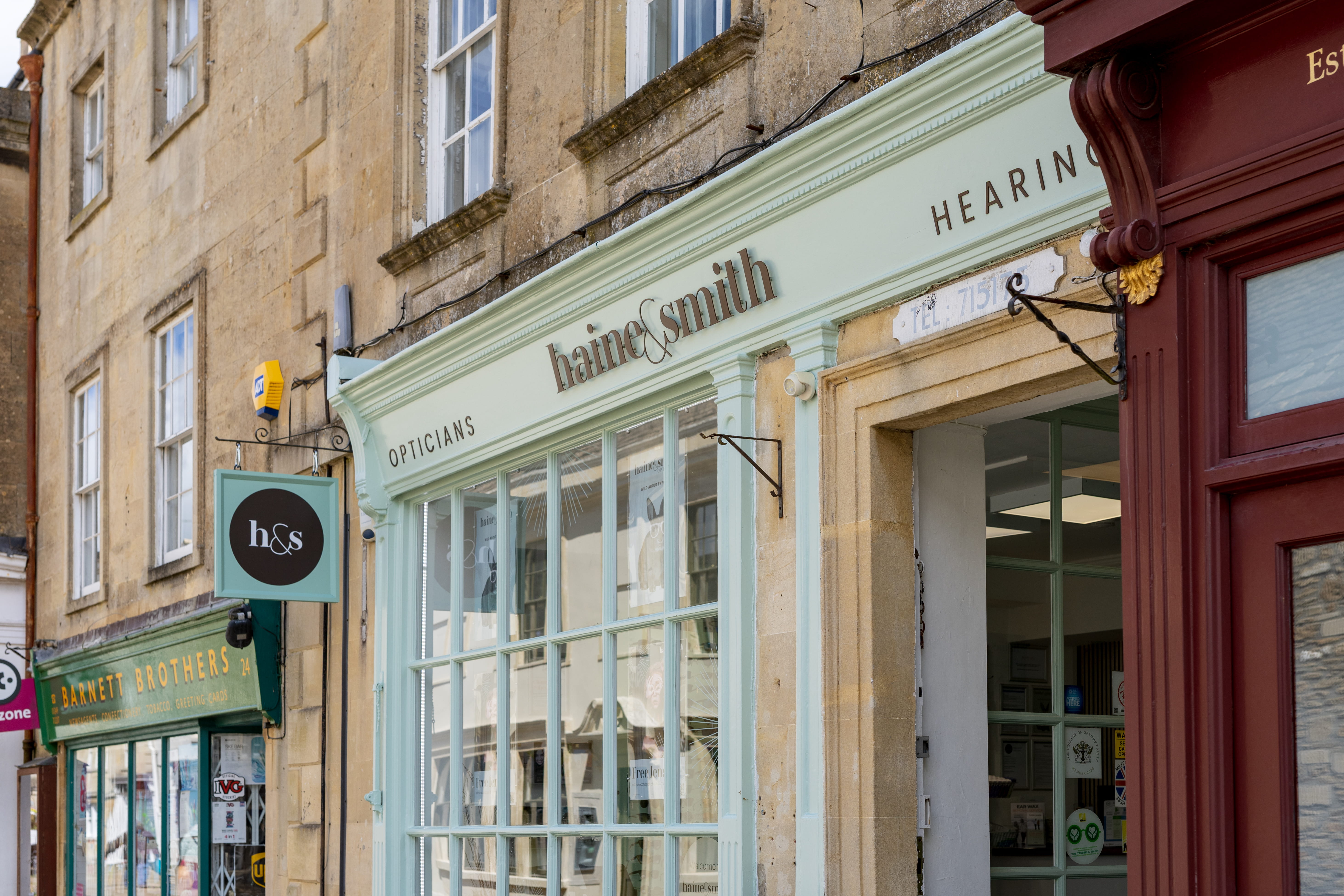 Exterior of a Haine & Smith Opticians and Hearing Care practice in a historic stone-fronted building, featuring large display windows, a hanging branded sign, and a welcoming entrance