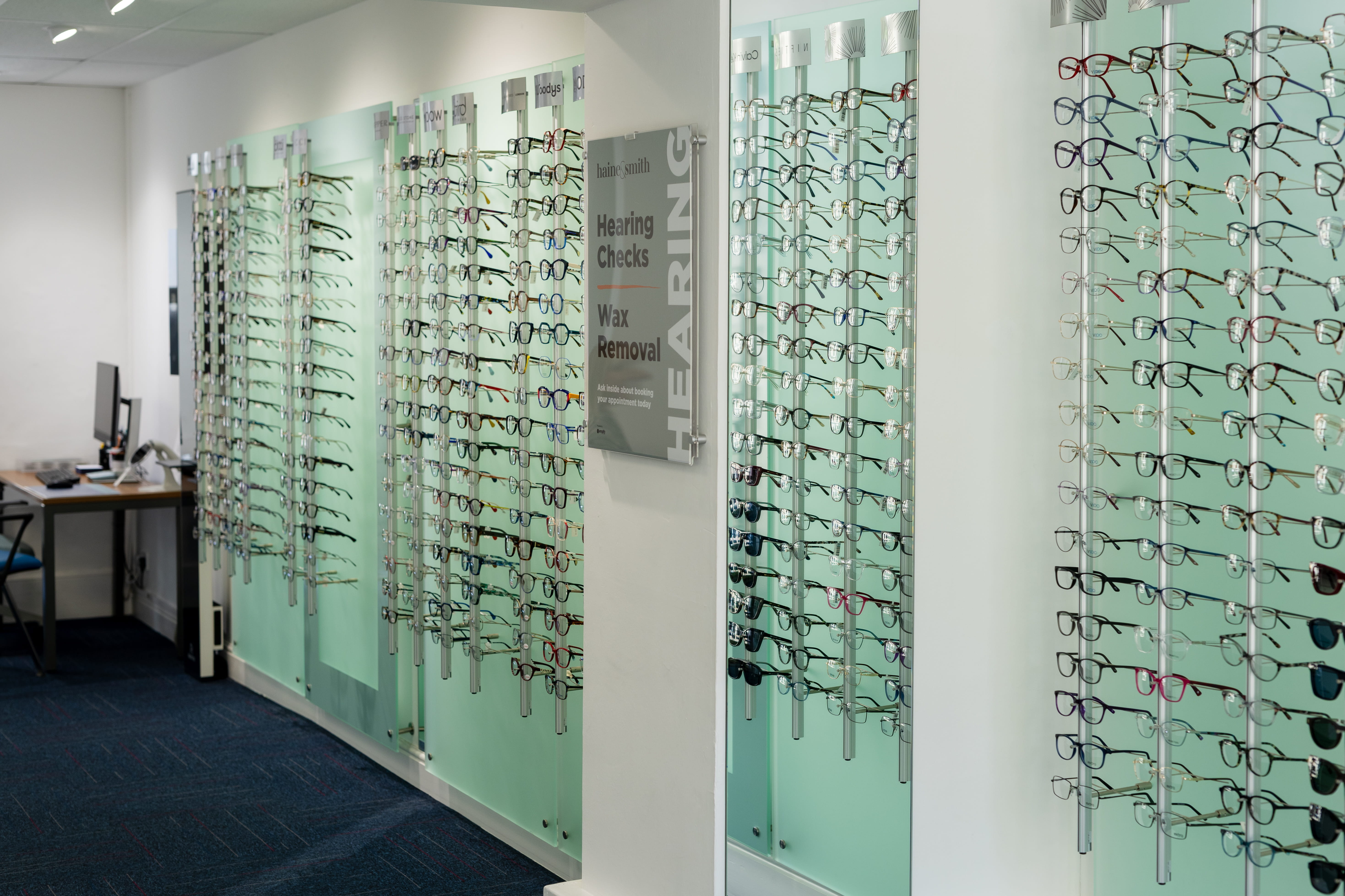 Eyewear display area inside a Haine & Smith Opticians practice featuring multiple wall-mounted glass panels showcasing a wide range of prescription glasses and sunglasses, with signage for hearing checks and ear wax removal services