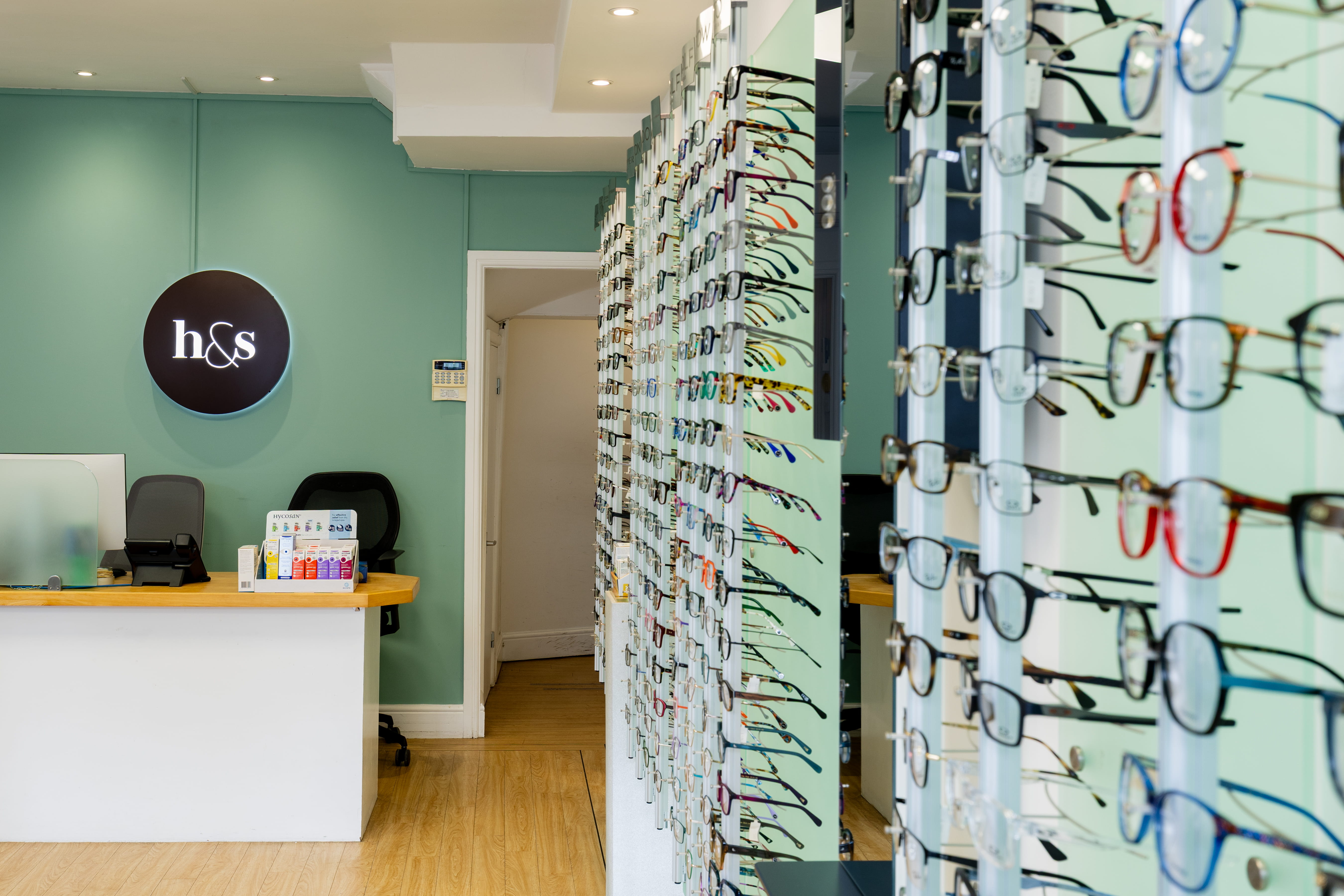 Contemporary optical practice interior with a reception desk and illuminated Haine & Smith logo, alongside extensive displays of prescription glasses and designer eyewear frames