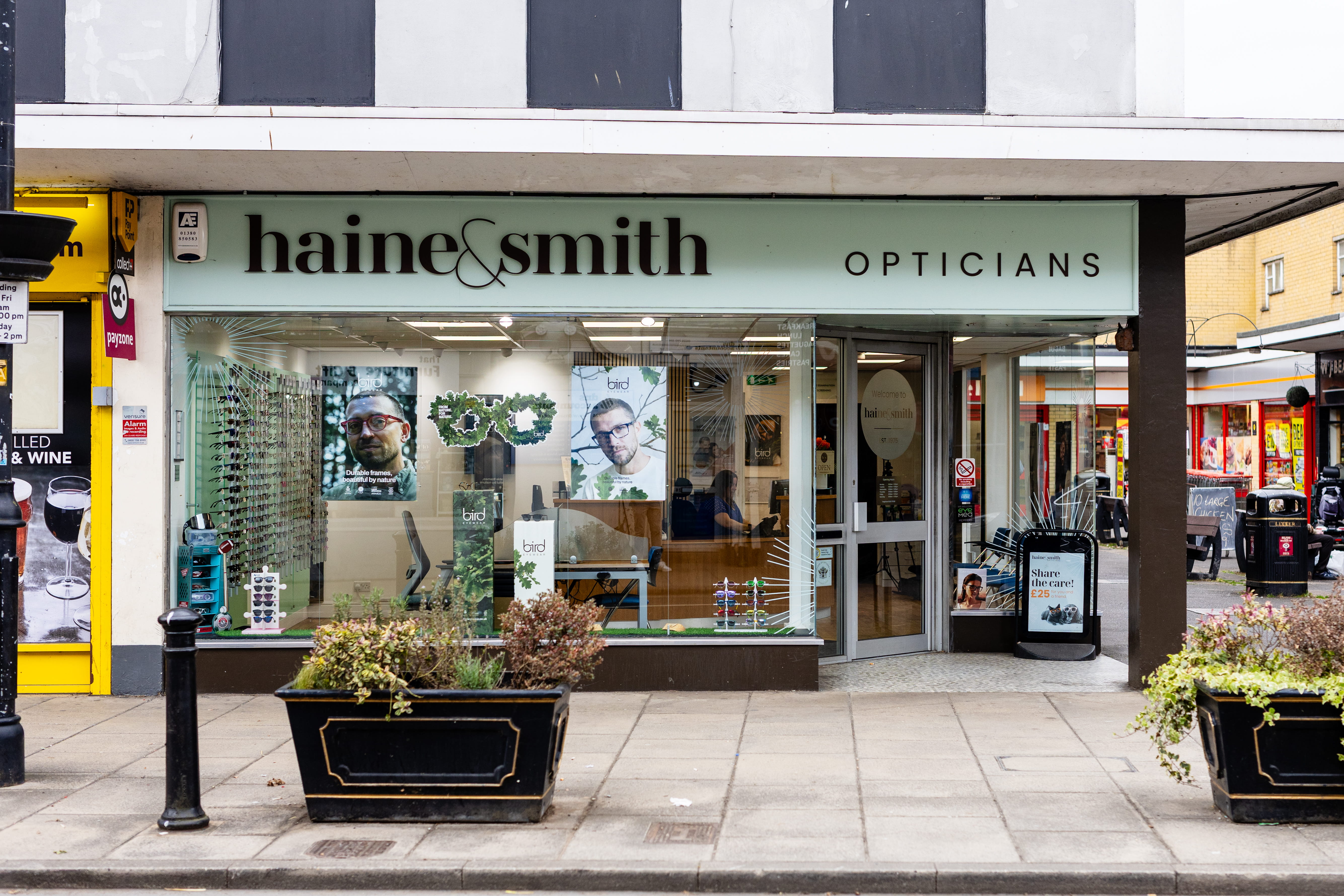 Exterior view of a Haine & Smith Opticians practice on a busy high street, featuring large display windows showcasing eyewear collections and eye care products