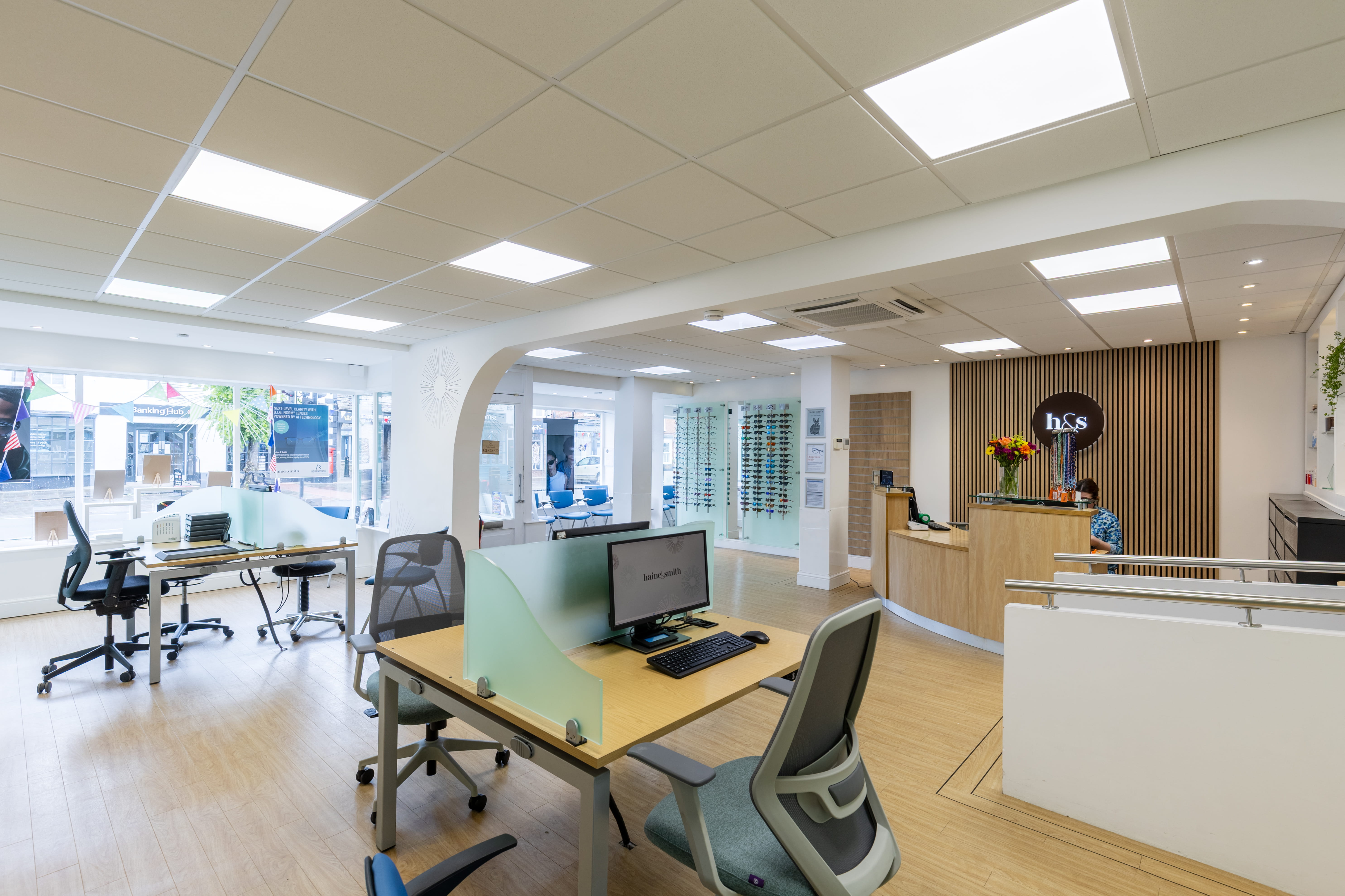Modern optical practice interior with consultation desks, designer eyewear displays mounted on glass panels, customer seating, and a bright, spacious showroom environment
