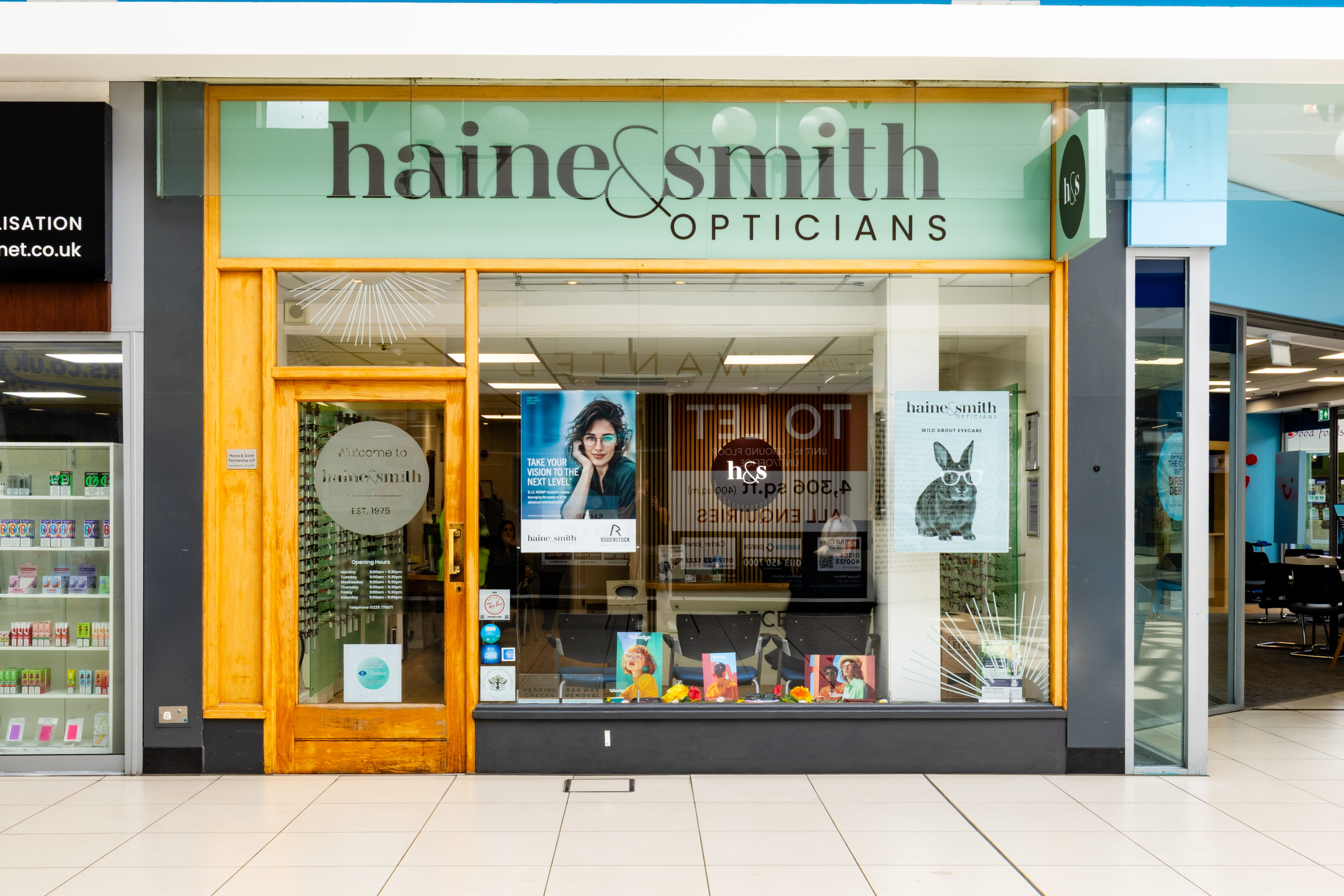 Front view of a Haine & Smith Opticians practice inside a shopping centre, featuring large display windows, eyewear posters, and a welcoming optical retail space