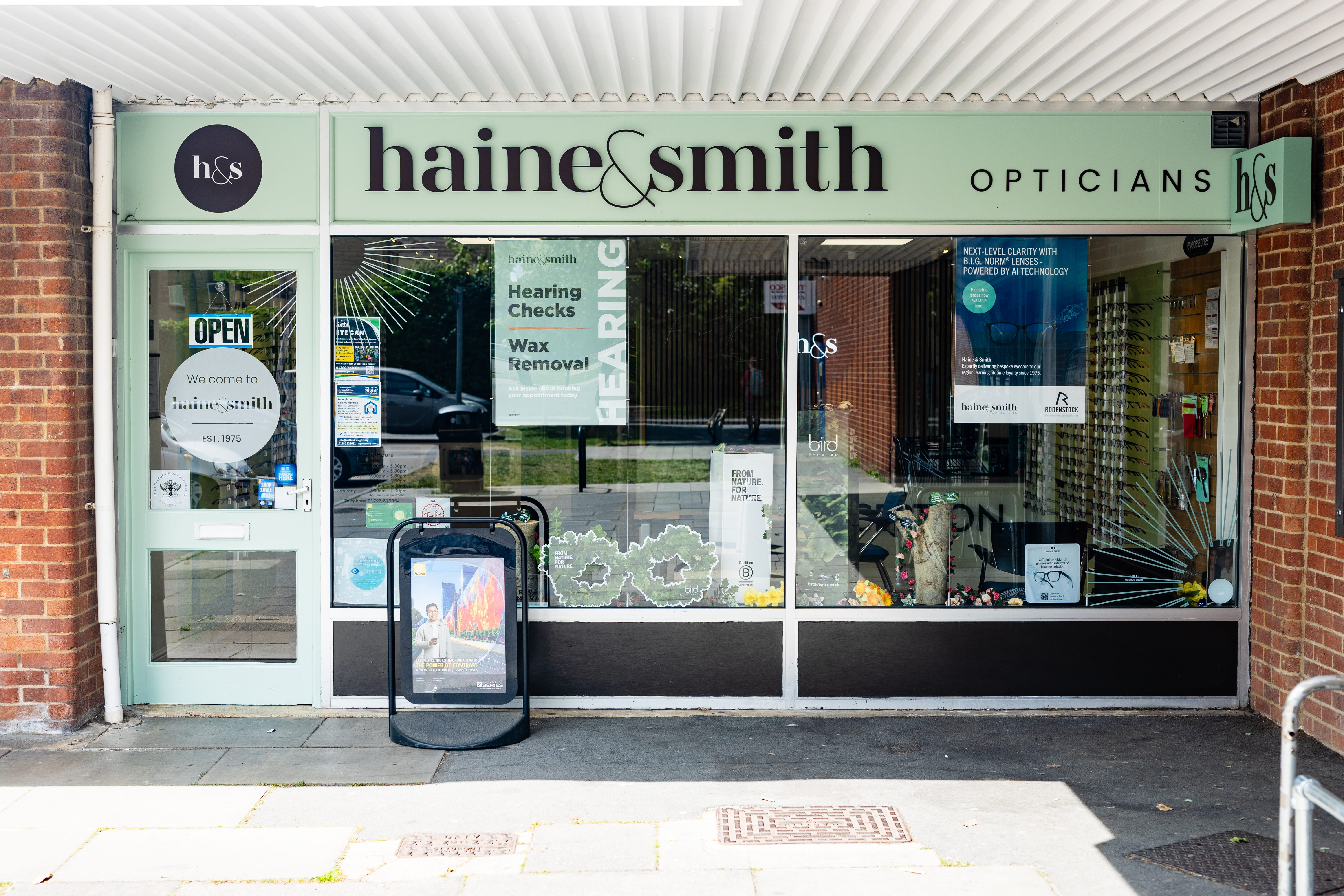 The exterior of Haine & Smith Opticians, a retail shopfront with a sage green fascia displaying the brand name and logo. The large glass window showcases promotional signage for hearing checks, wax removal, and AI-powered lenses, along with eyewear display