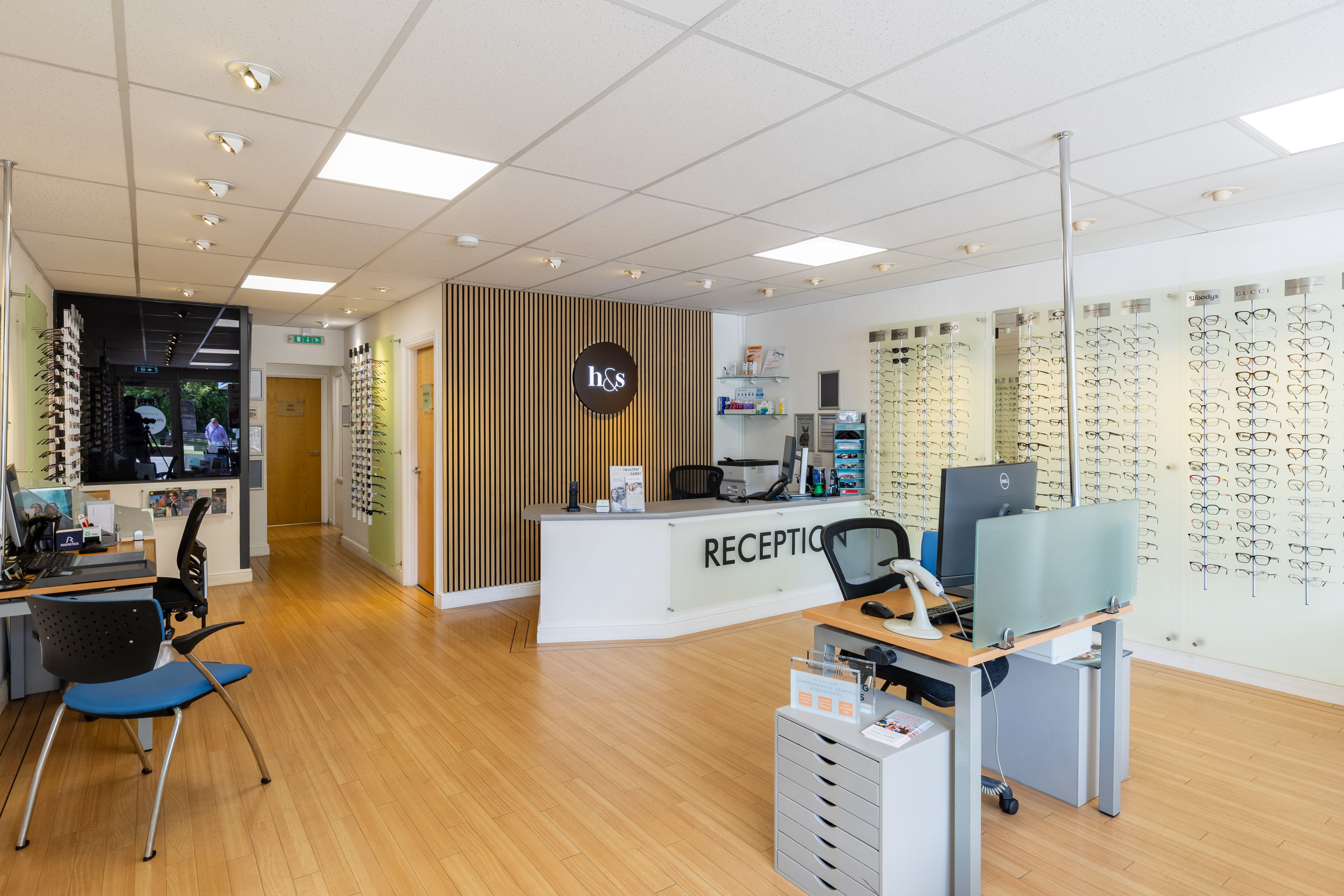 The bright and modern interior of Haine & Smith Opticians, featuring a curved white reception desk with the "h&s" logo mounted on a natural wood slat accent wall behind it. The spacious showroom has light wood flooring, recessed ceiling lighting, and walls