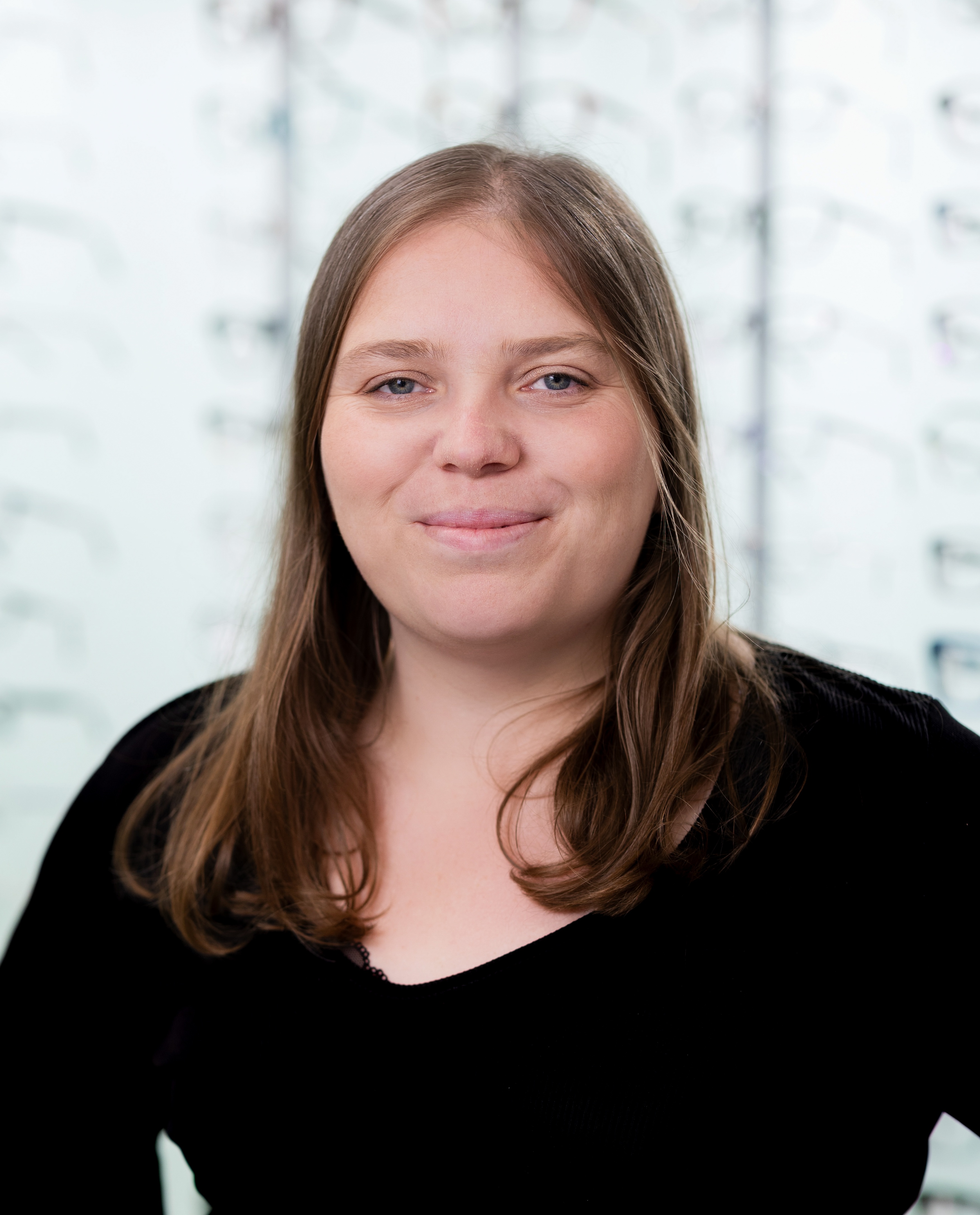 Smiling optician wearing round-frame glasses standing in front of a display wall showcasing a wide selection of prescription eyewear frames