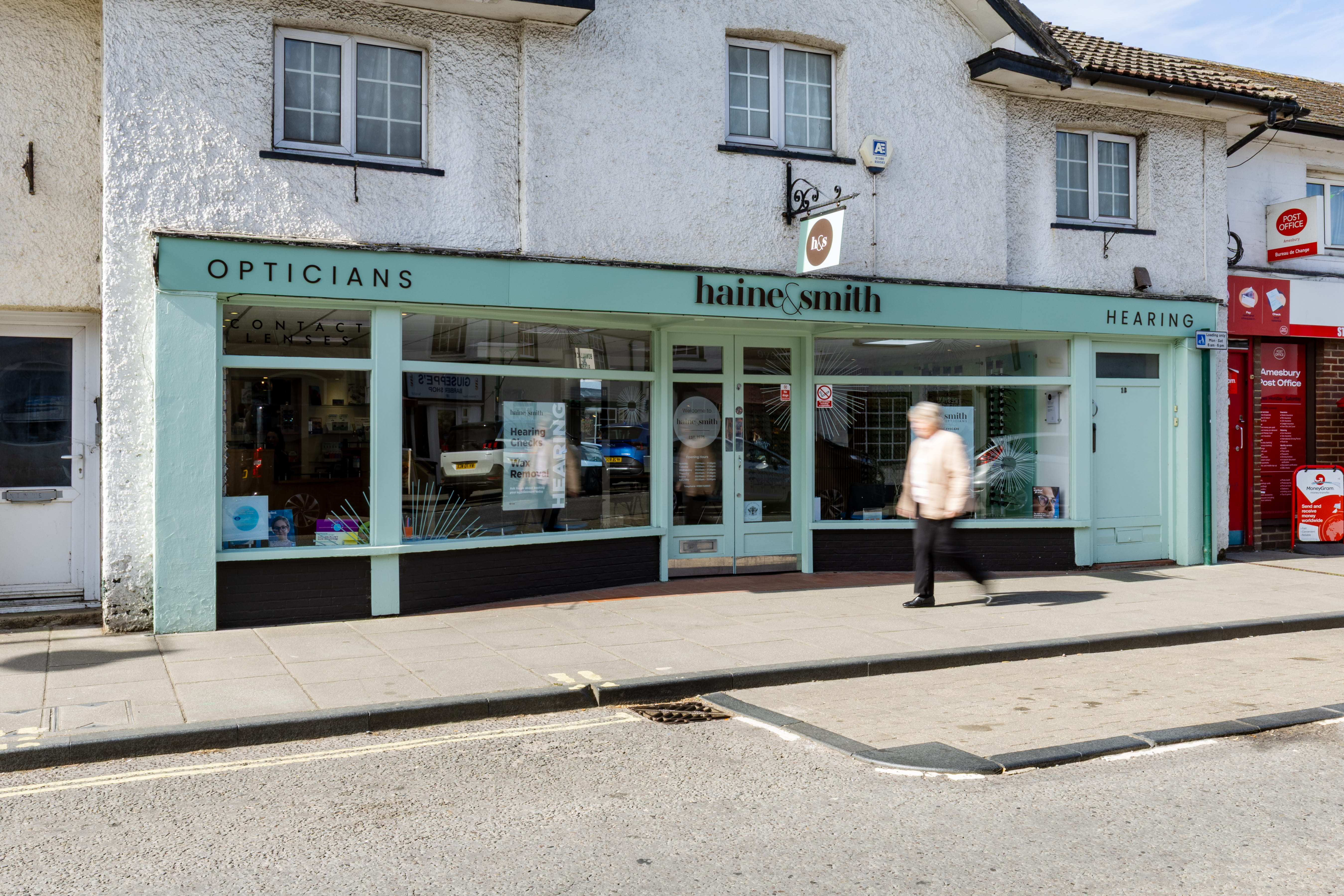 Exterior of a Haine & Smith Opticians and Hearing Care practice on a high street, featuring large display windows, contact lens services, and a pedestrian walking past the storefront