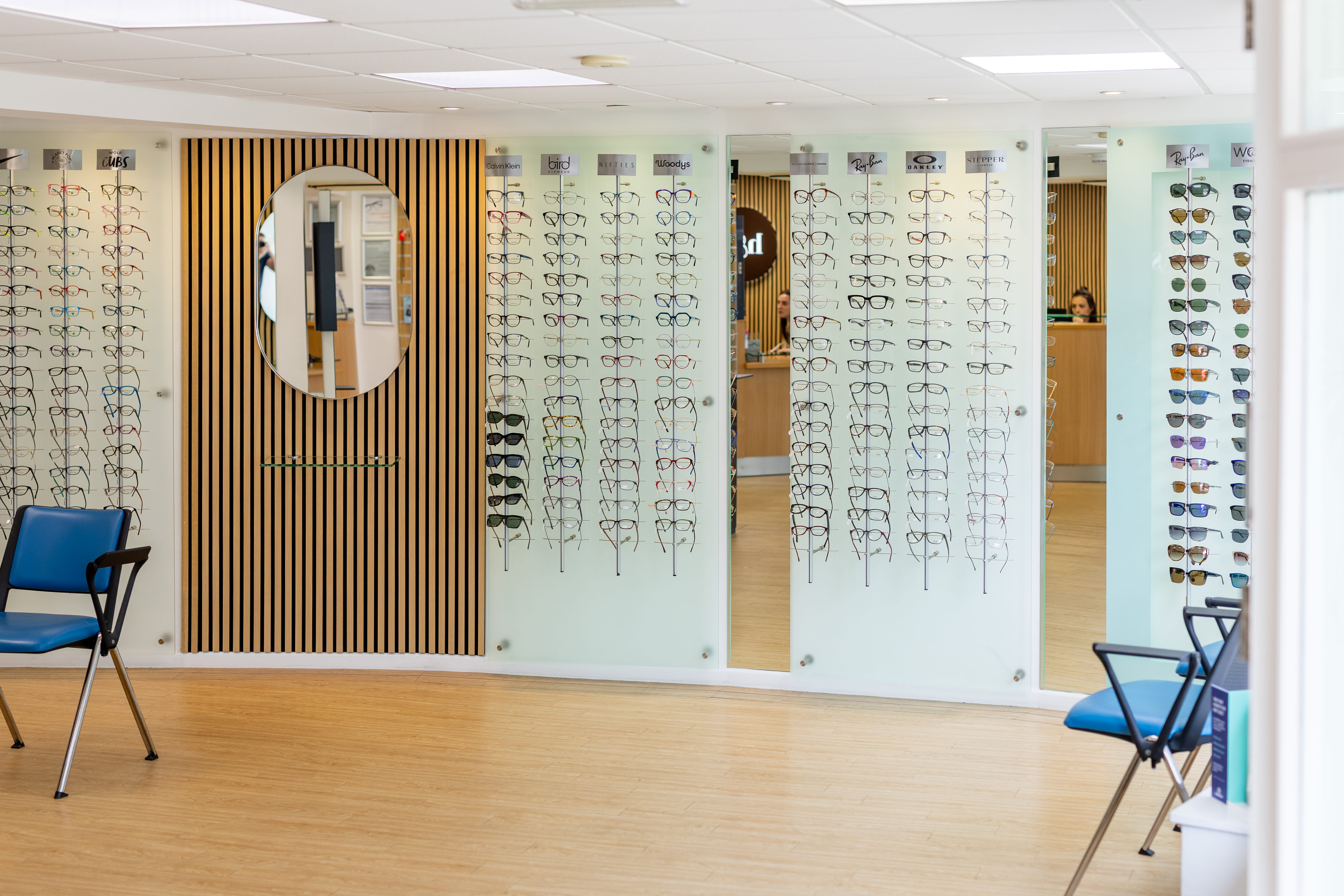 Bright optical showroom with wall-mounted displays of prescription glasses and sunglasses, blue seating, wooden flooring, and a modern eyewear consultation area
