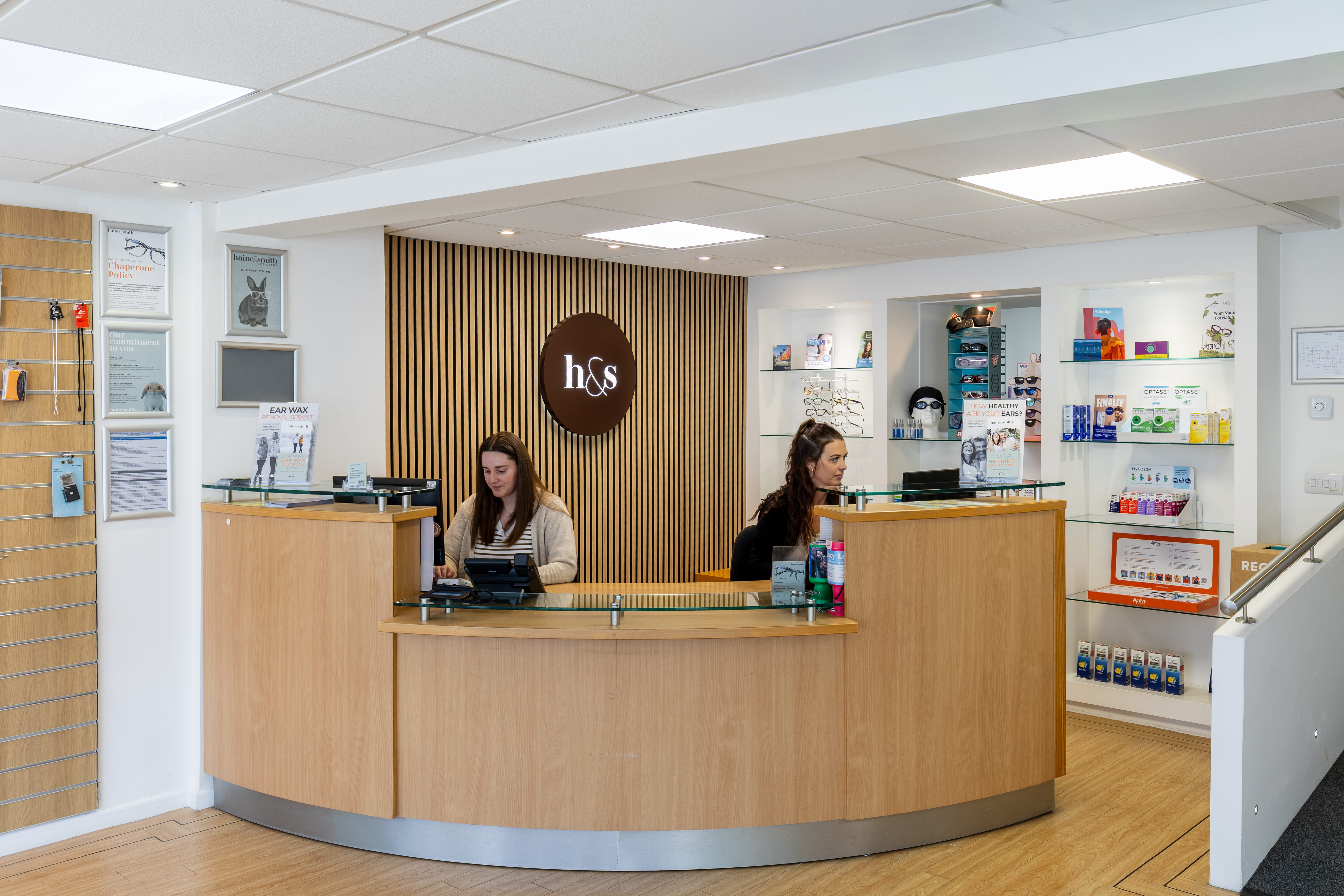 Reception area inside a Haine & Smith Opticians practice, featuring a curved wooden front desk, team members assisting customers, product displays, and a modern eyewear retail environment