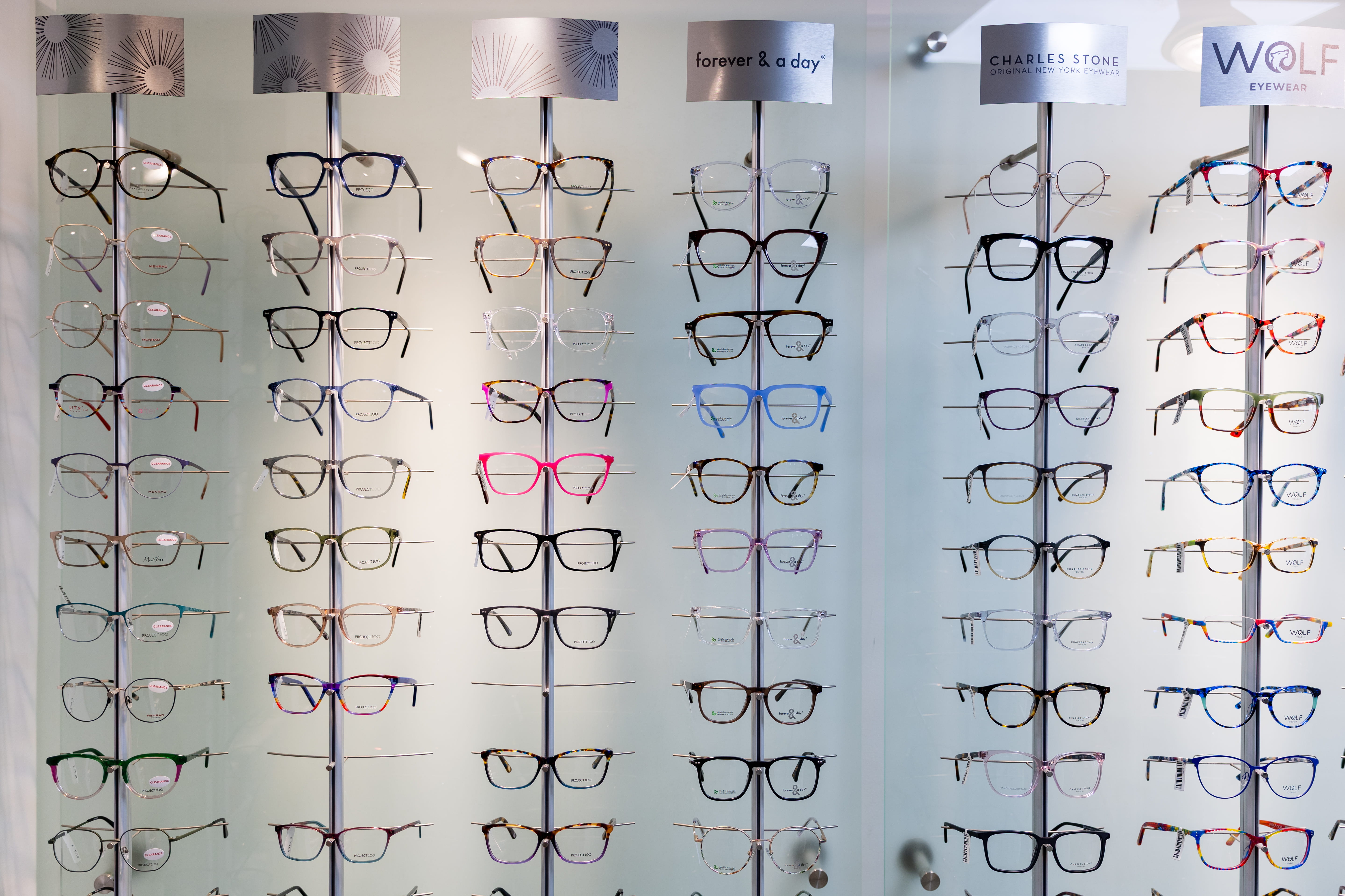 Display of designer prescription eyeglass frames arranged on illuminated glass panels, featuring a wide selection of styles, colours, and brands within a modern opticians showroom