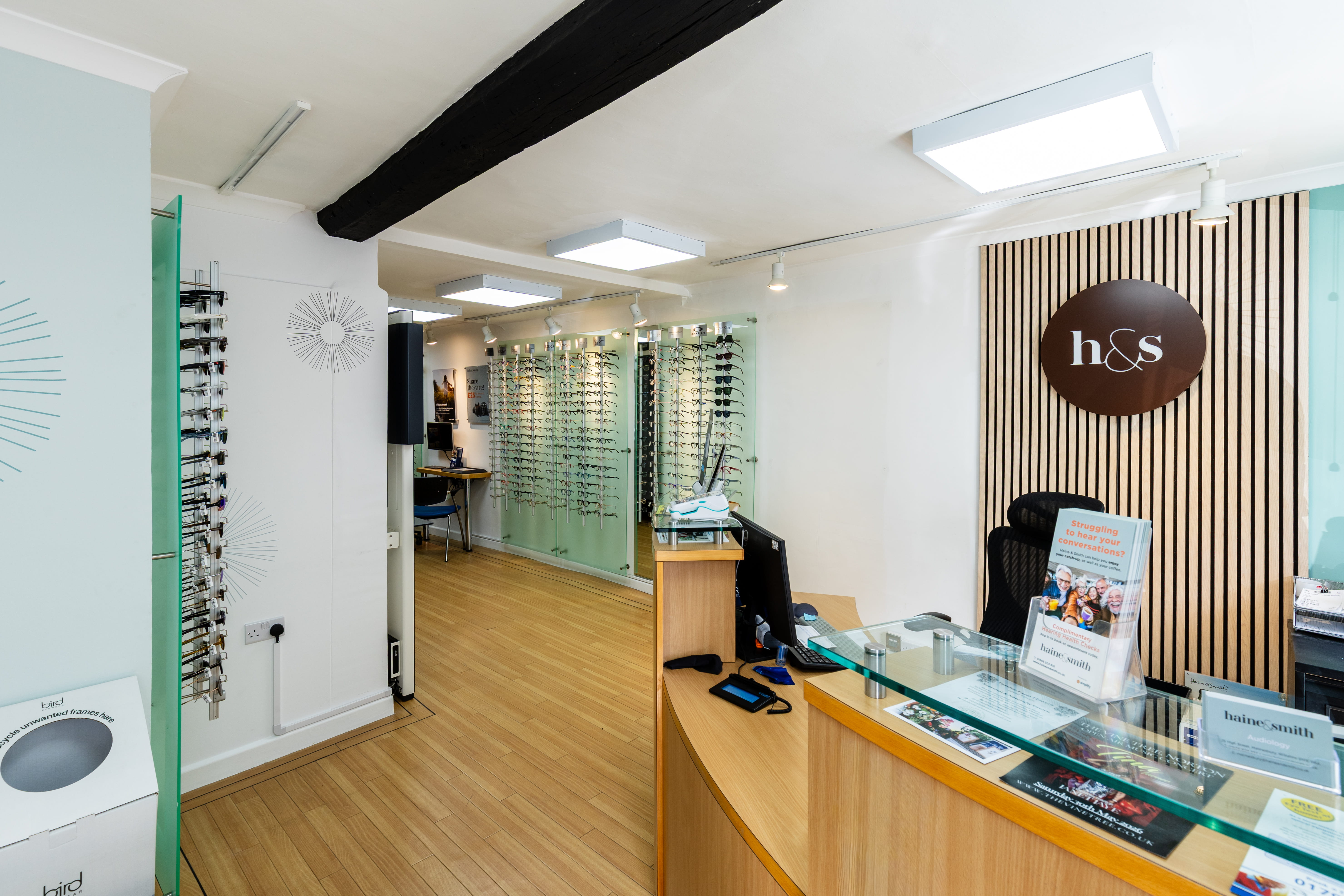 Welcoming reception area inside a Haine & Smith Opticians practice, featuring a wooden reception desk, illuminated brand logo, eyewear displays, and a bright showroom with modern fittings