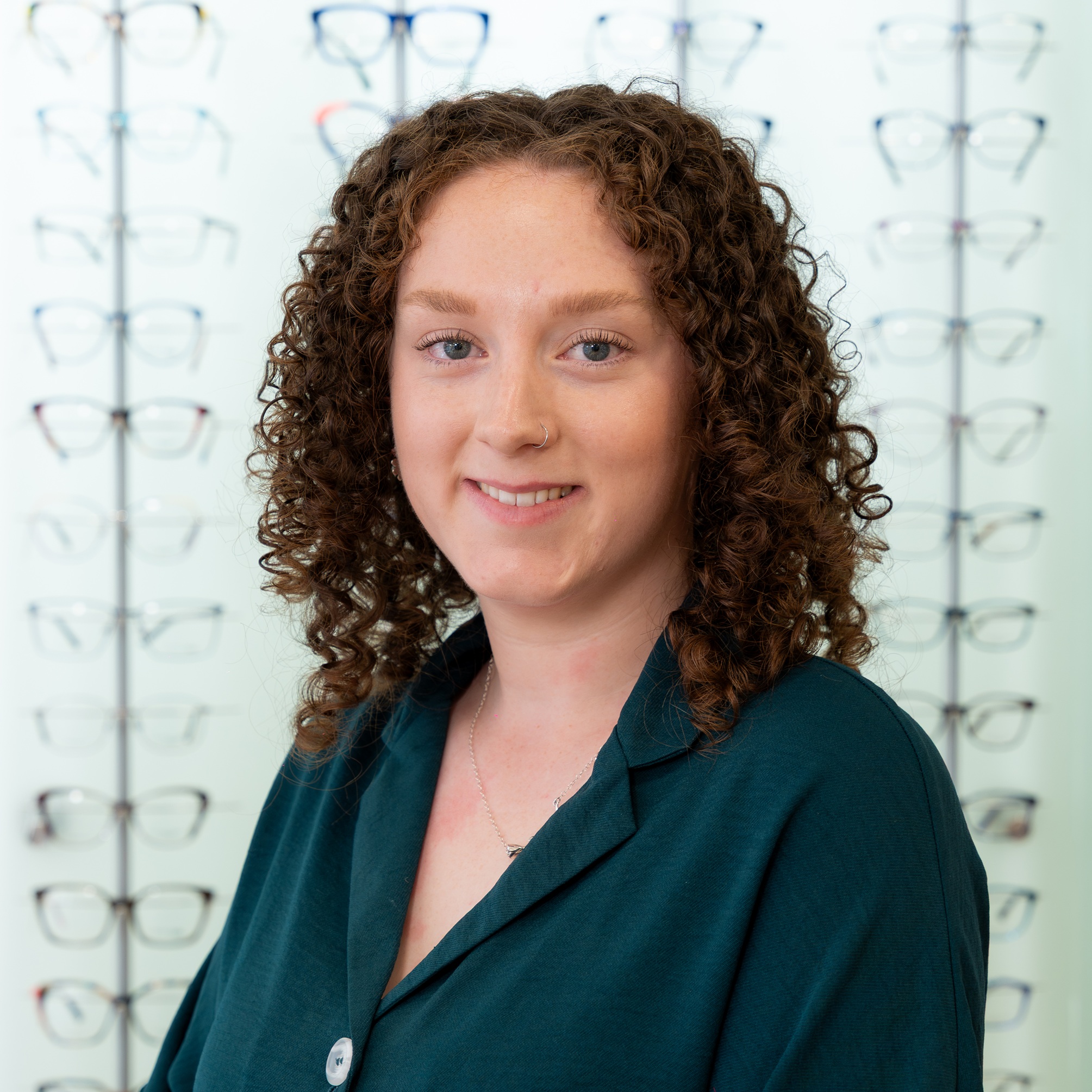Smiling optician wearing round-frame glasses standing in front of a display wall showcasing a wide selection of prescription eyewear frames