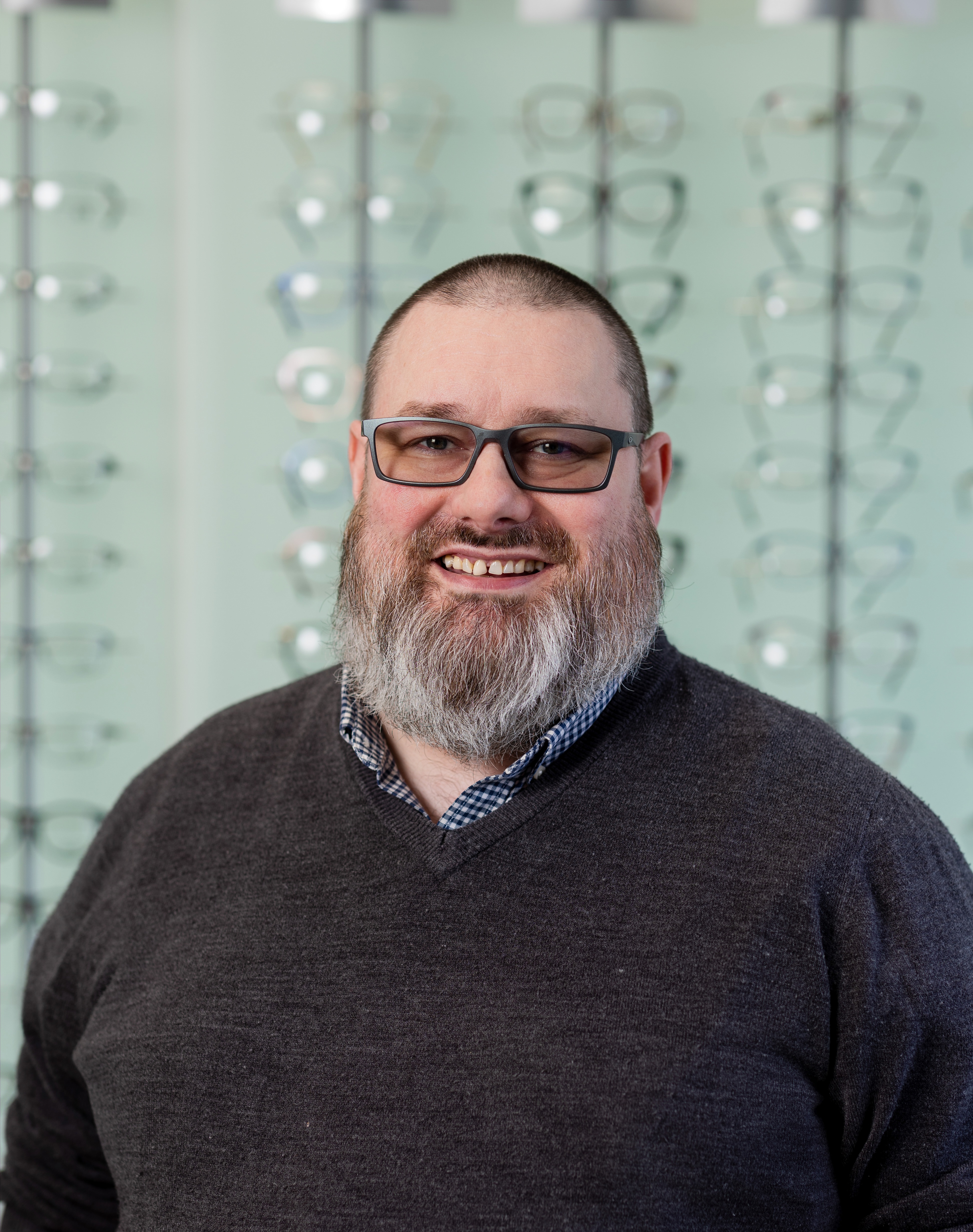 Smiling optician wearing round-frame glasses standing in front of a display wall showcasing a wide selection of prescription eyewear frames