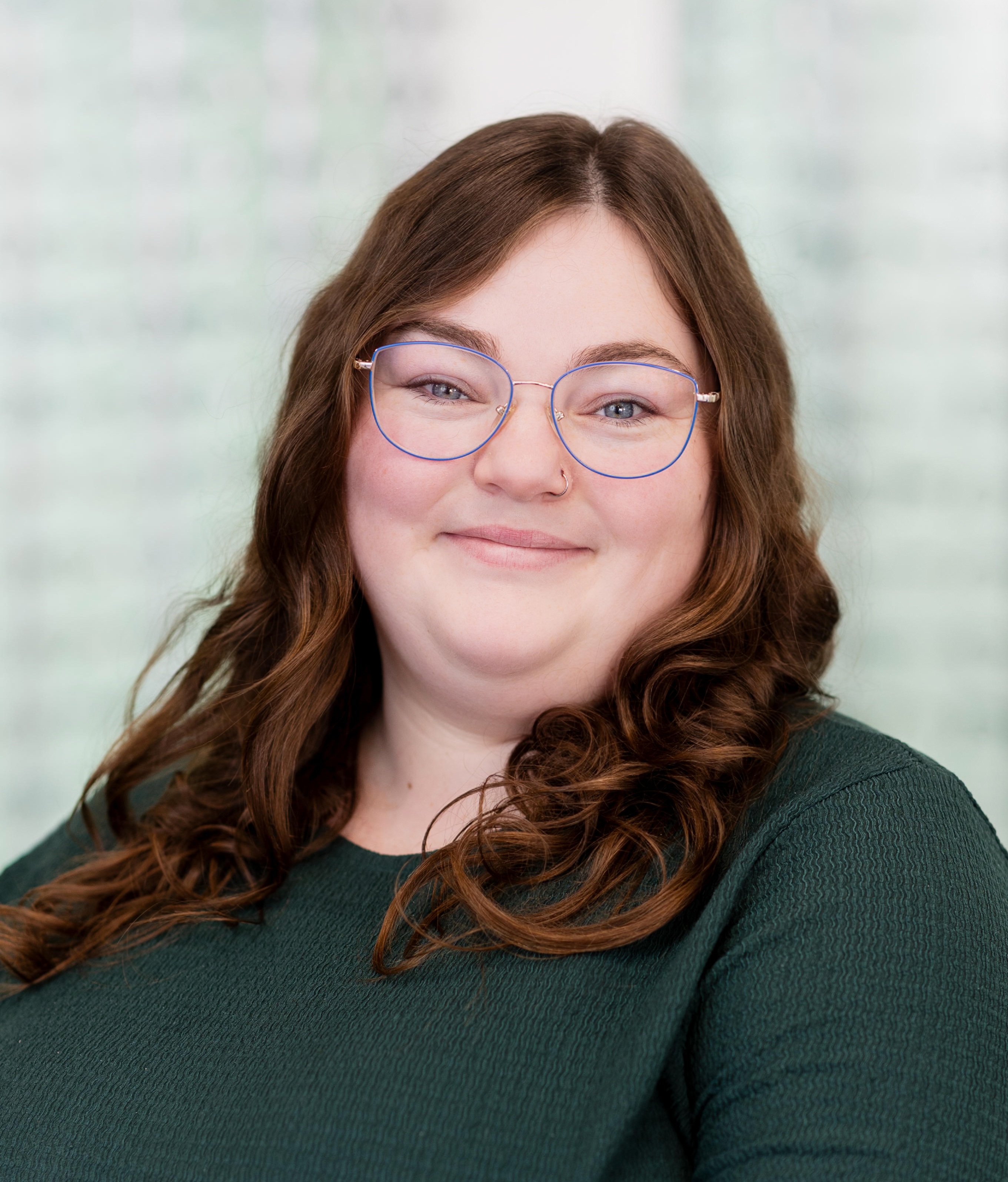 Smiling optician wearing round-frame glasses standing in front of a display wall showcasing a wide selection of prescription eyewear frames