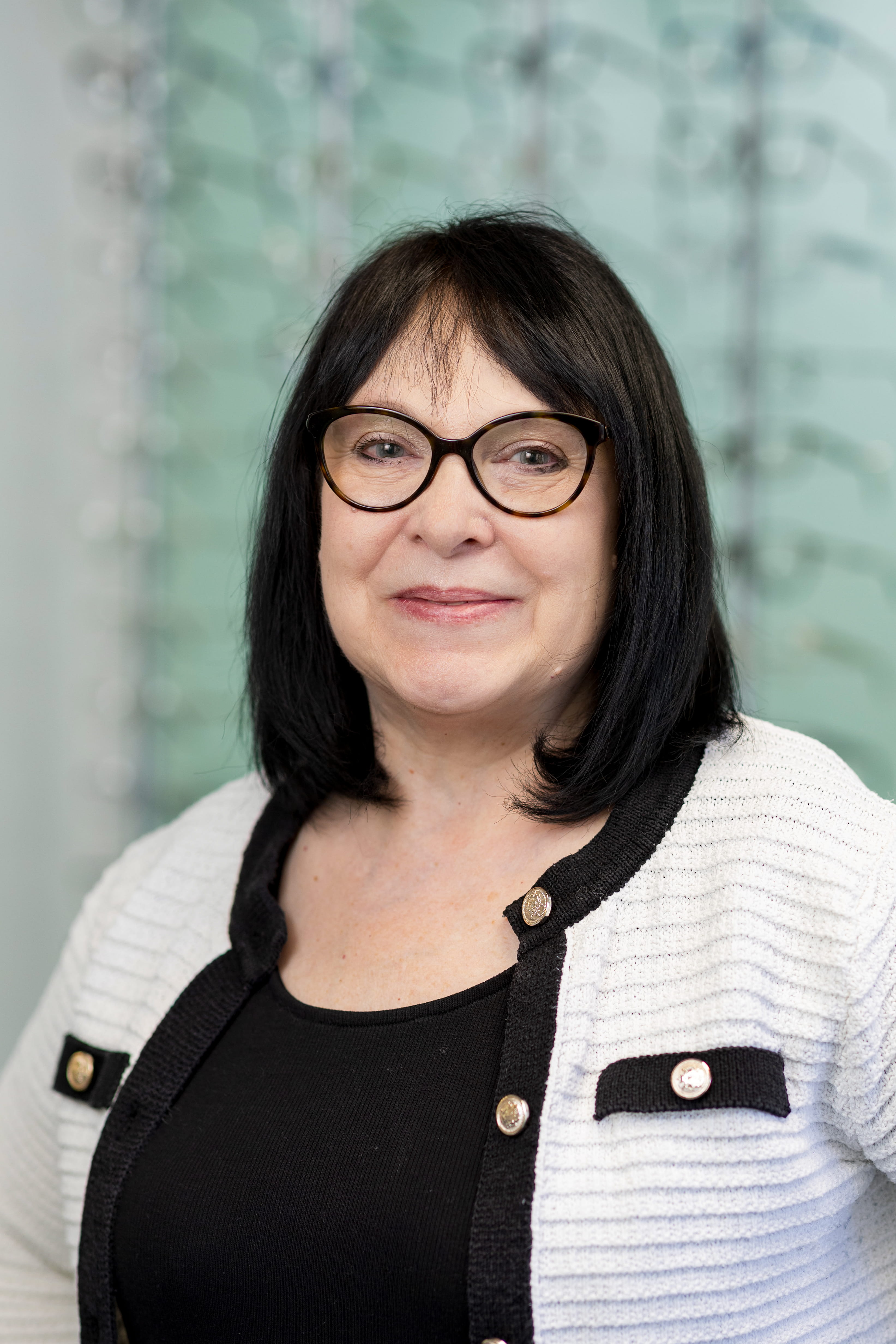 Smiling optician wearing round-frame glasses standing in front of a display wall showcasing a wide selection of prescription eyewear frames