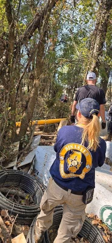 People walking through a wooded area with supplies like coiled black tubing and bags, one person wearing a navy shirt with a rescue emblem.