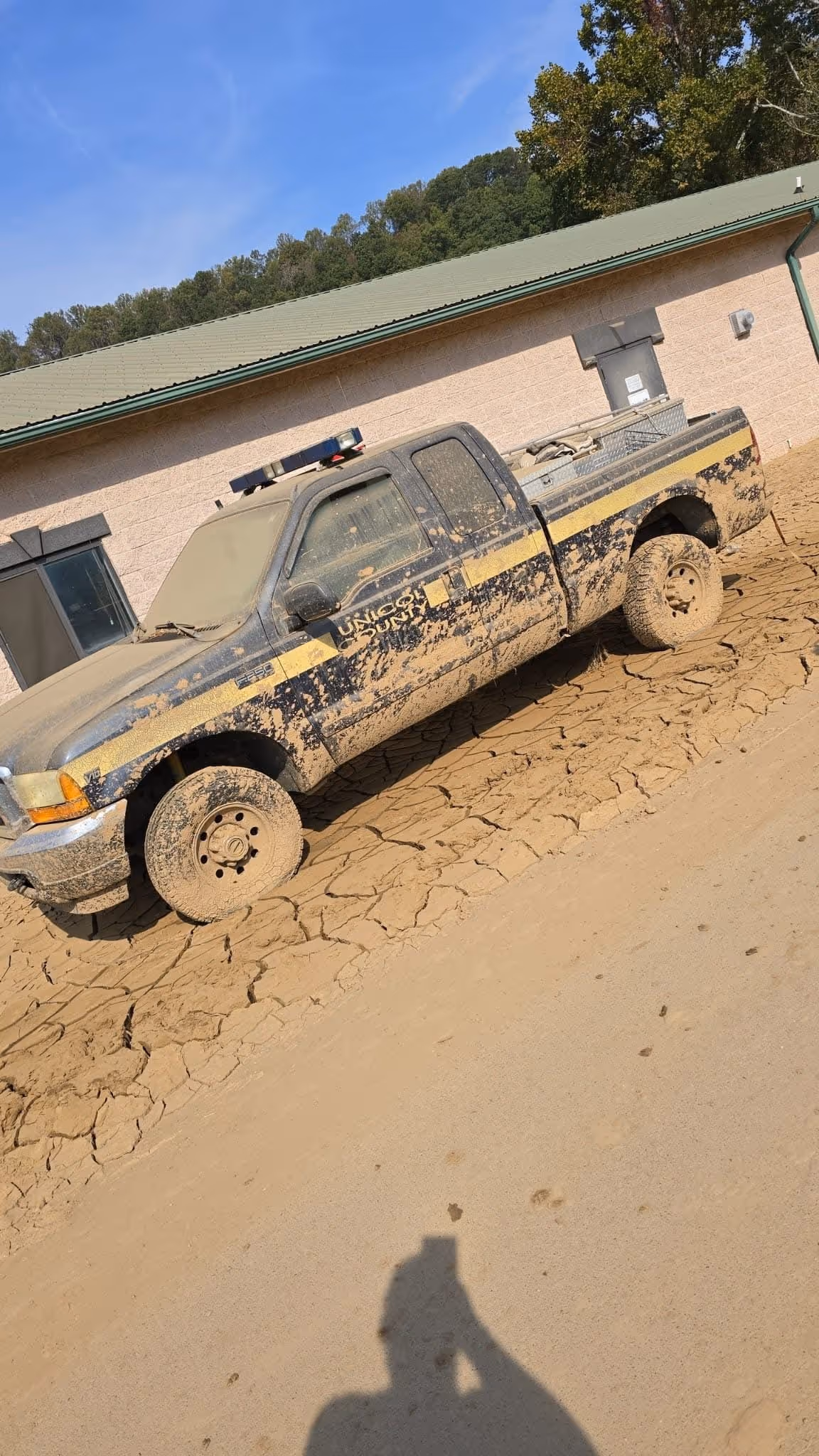 Mud-covered utility pickup truck with a light bar on the roof parked on cracked dry mud in front of a building.