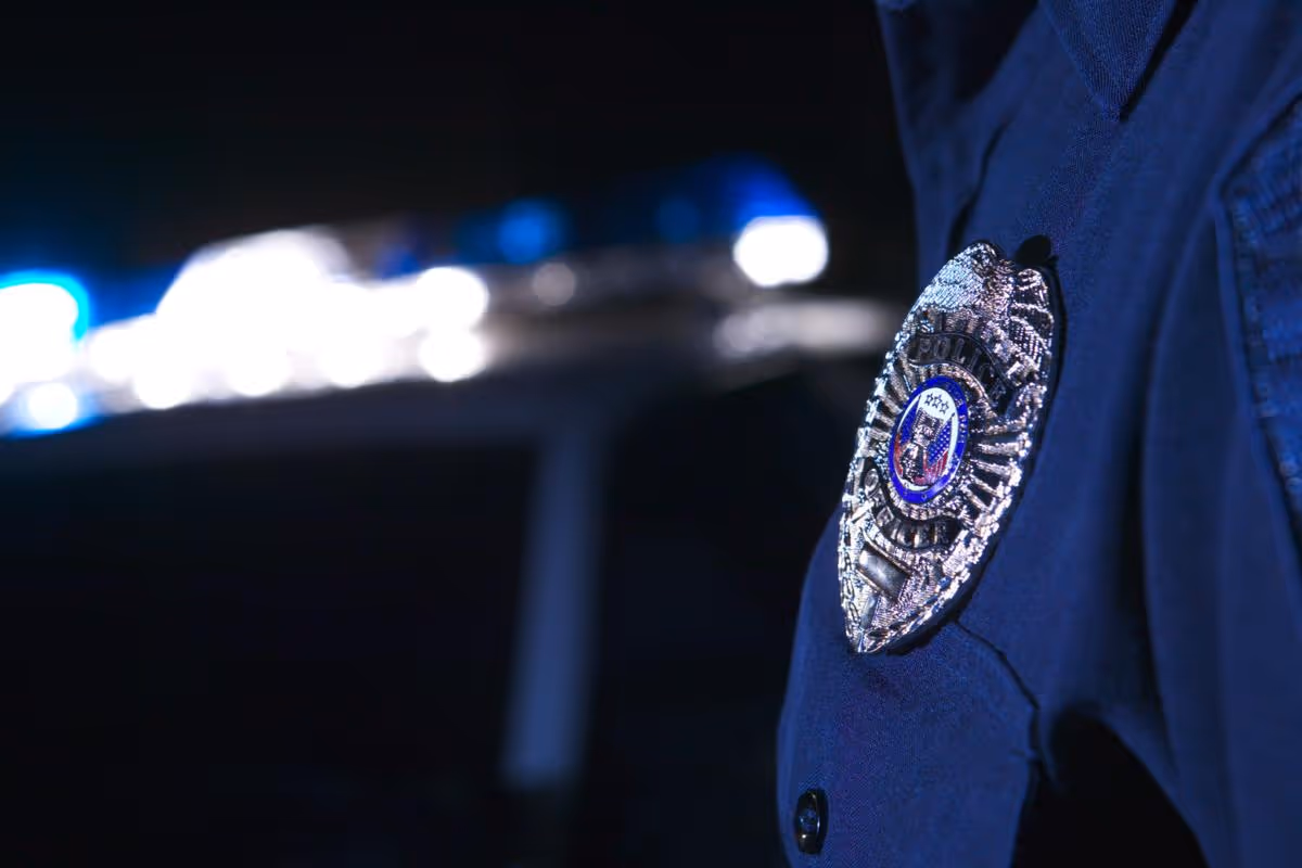 Close-up of a police officer's badge on a dark uniform with blurred police car lights in the background.