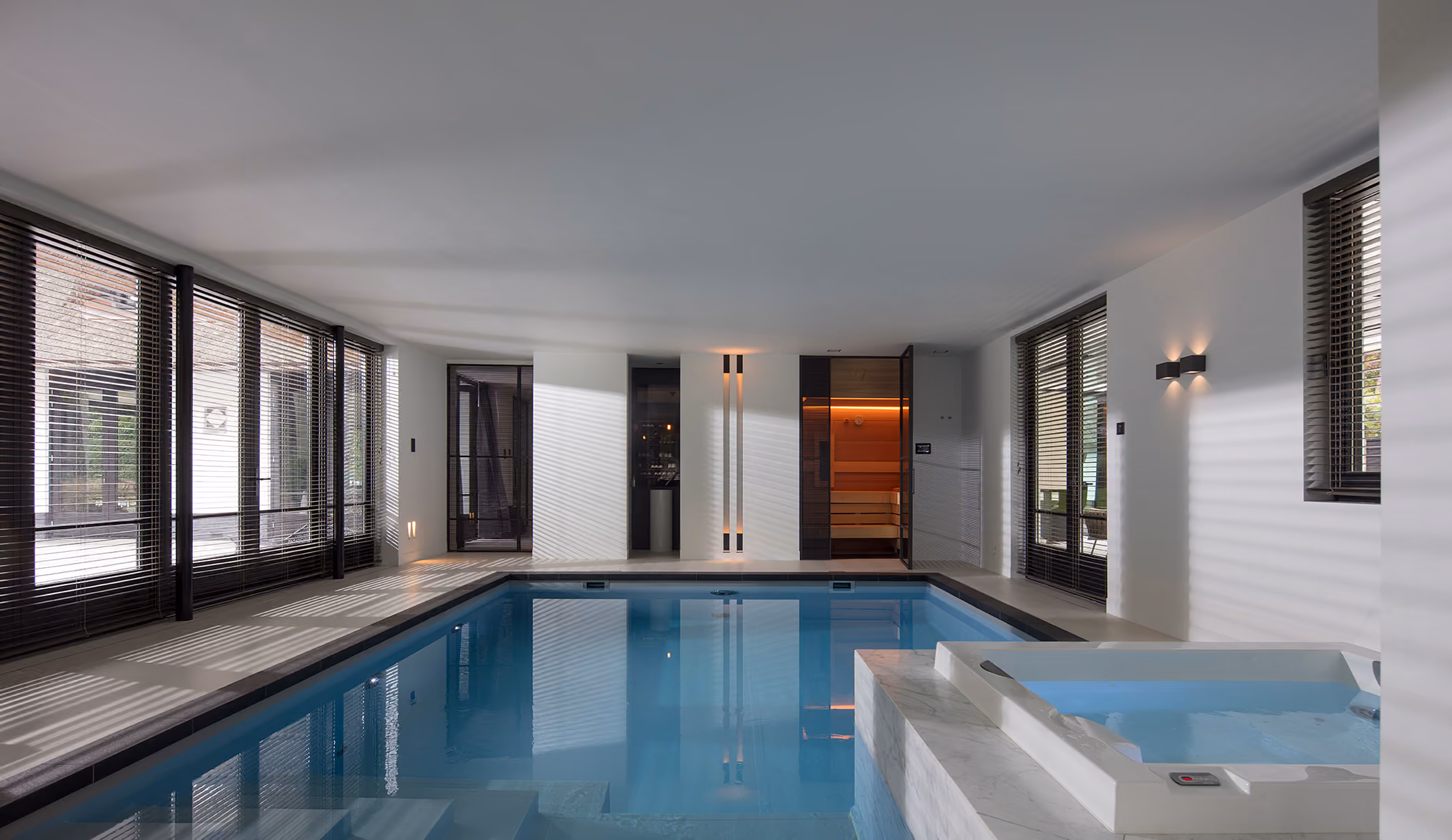 Binnenzwembad met blauwe tegels en jacuzzi in moderne kamer met ramen en houten jaloezieën.