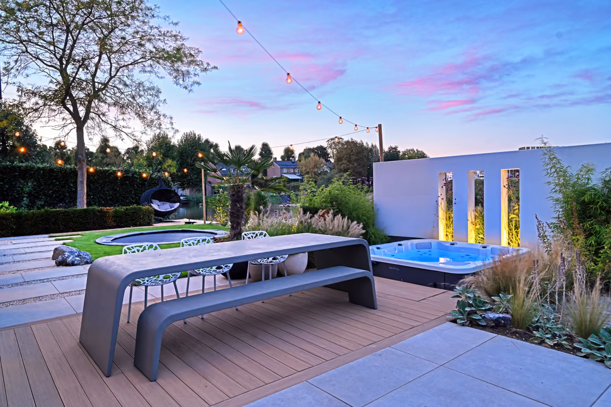 Moderne tuin met strakke picknicktafel, hangstoel, jacuzzi en sfeerverlichting bij zonsondergang.