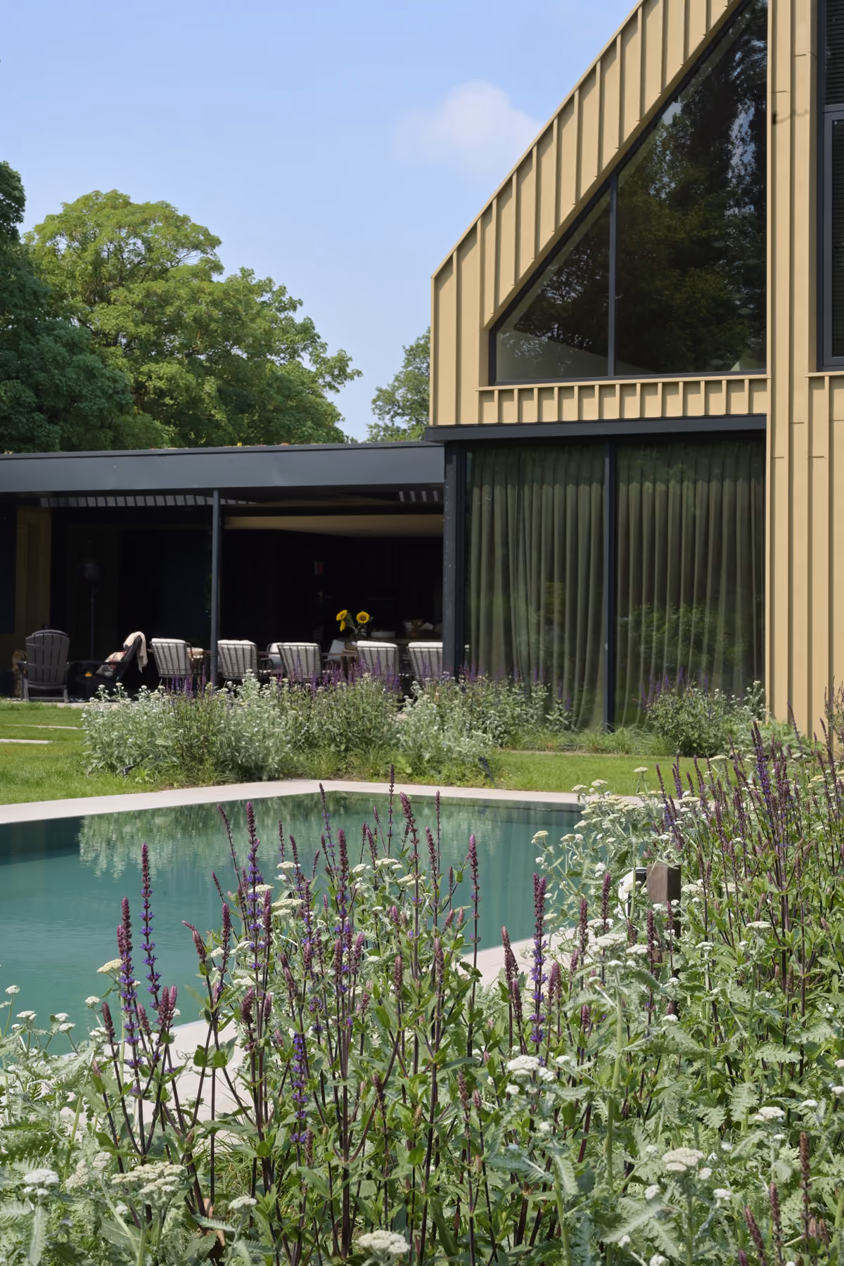 Moderne woning met groot raam, groen gazon, bloeiende planten en zwembad in de tuin.