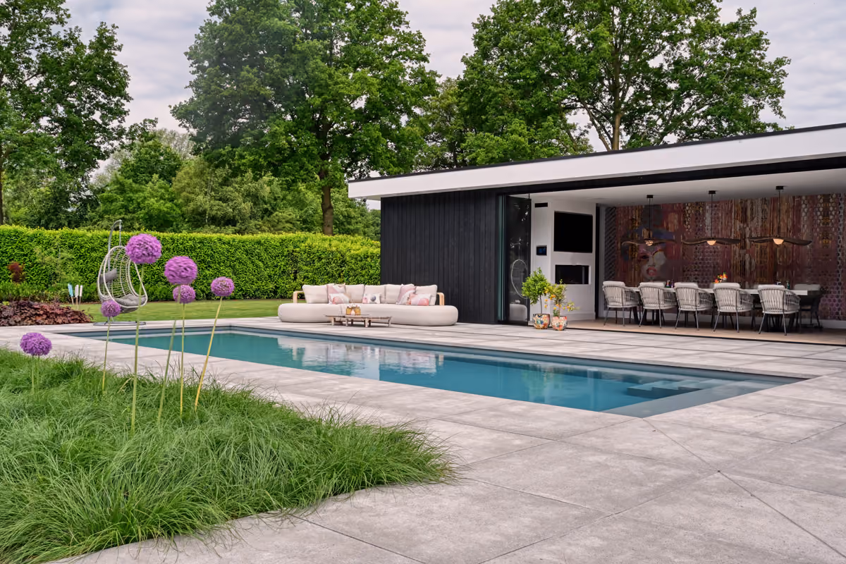 Moderne tuin met zwembad, loungebank, hangstoel en ruime overdekte eethoek met kunstmuur.