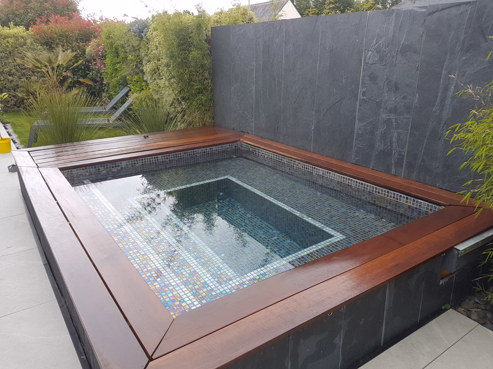 Moderne buitenjacuzzi met mozaïektegels en houten rand, omgeven door groene planten en een zwarte muur.