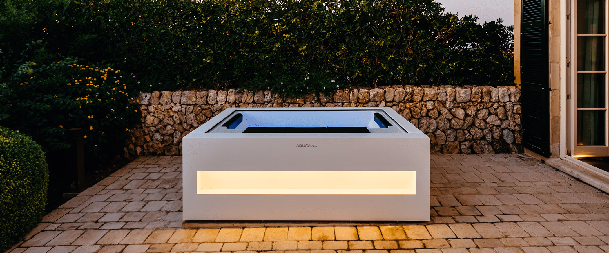 Witte moderne buitenjacuzzi met licht, stenen muur en groen in de achtergrond.
