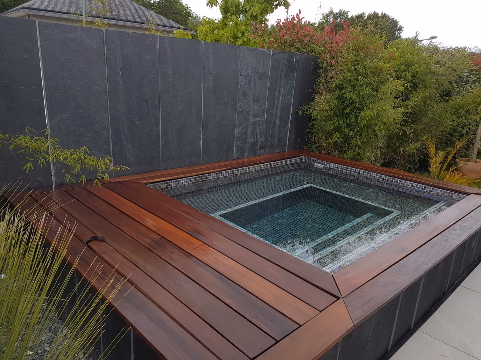 Moderne houten jacuzzi met glazen tegelafwerking in een tuin met groene planten.