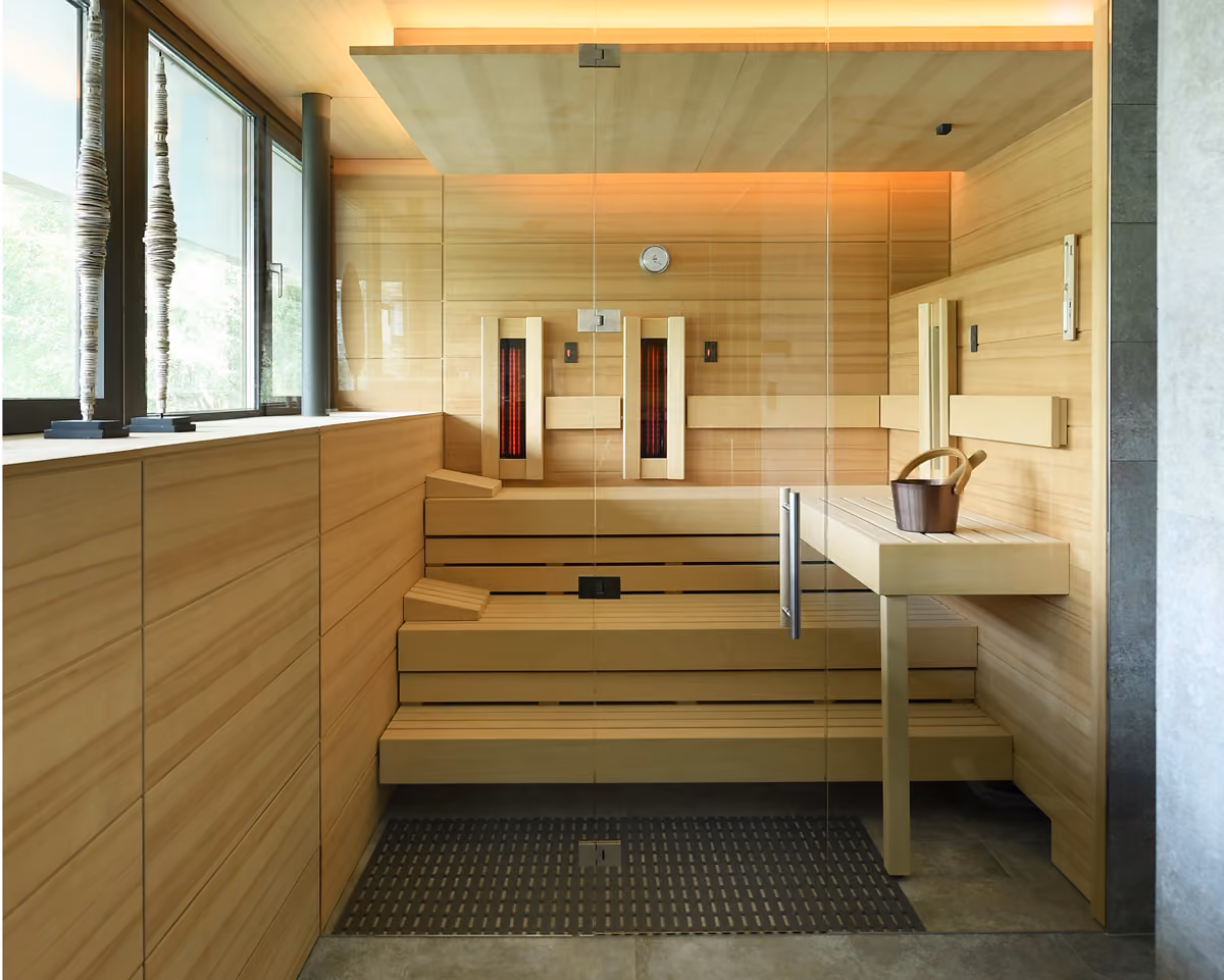 Moderne sauna met glazen deur, houten banken en een houten emmer op een plank.
