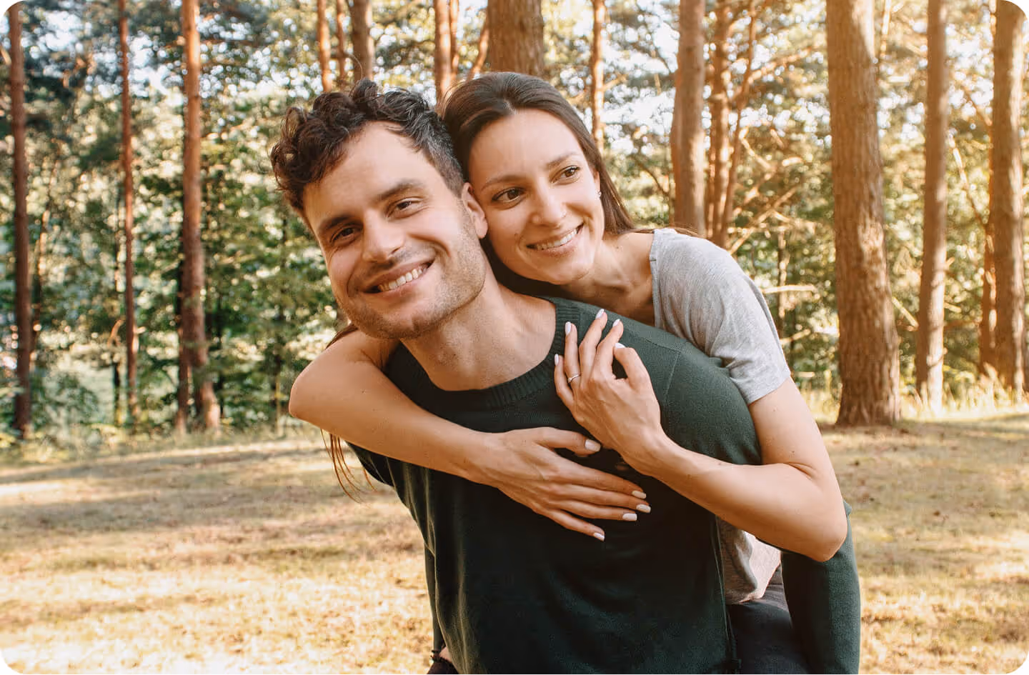 Smiling couple outdoors, with the woman playfully hugging the man from behind in a forest setting