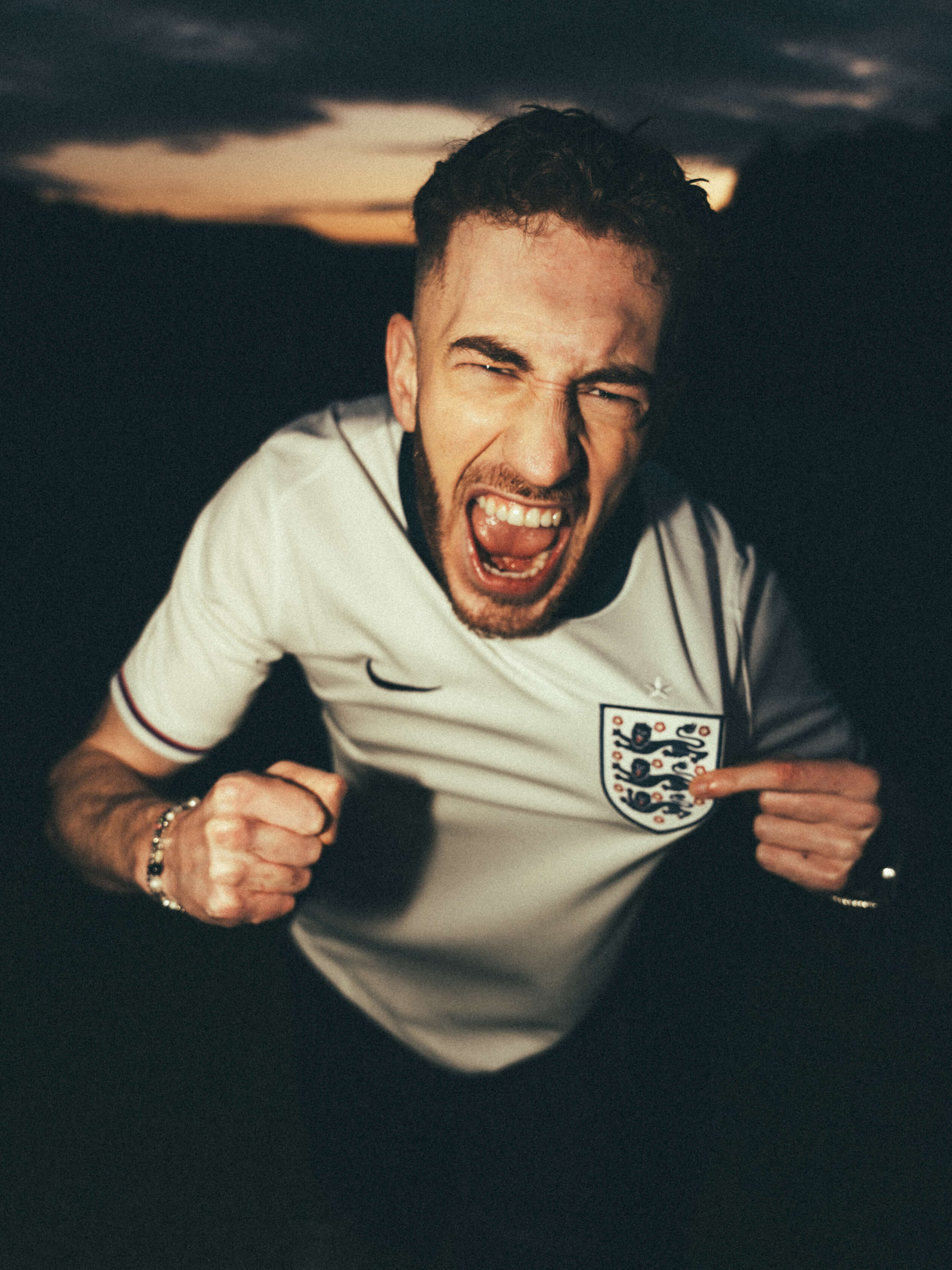 Singer LEWOUT poses in a England football shirt, holding the badge in his left hand and clenching his right—shouting as if he's scored a goal