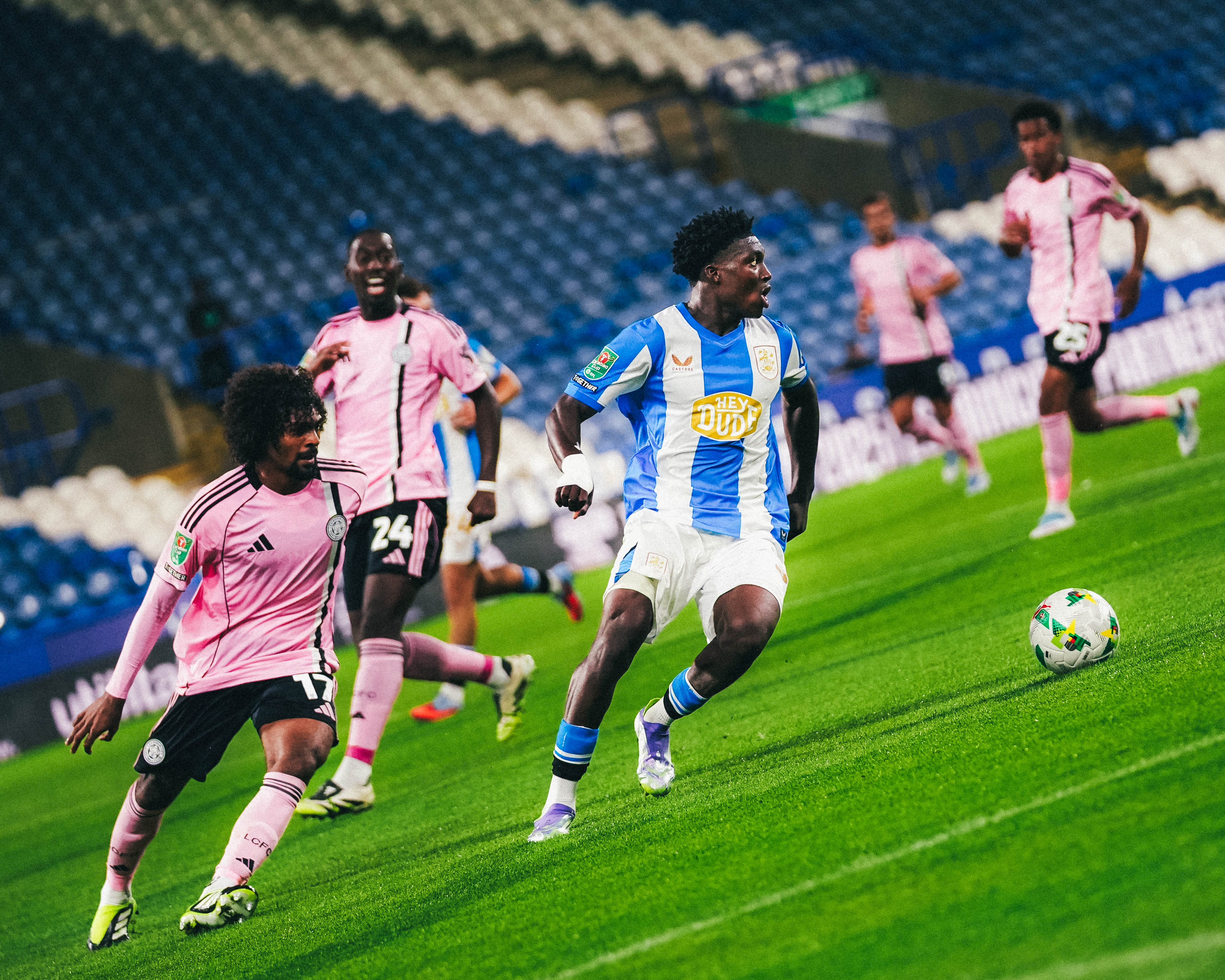 Cameron Ashia dribbling past a Leicester City defender for Huddersfield Town. Wearing a blue and white striped football shirt, and the Leicester players don an all pink kit