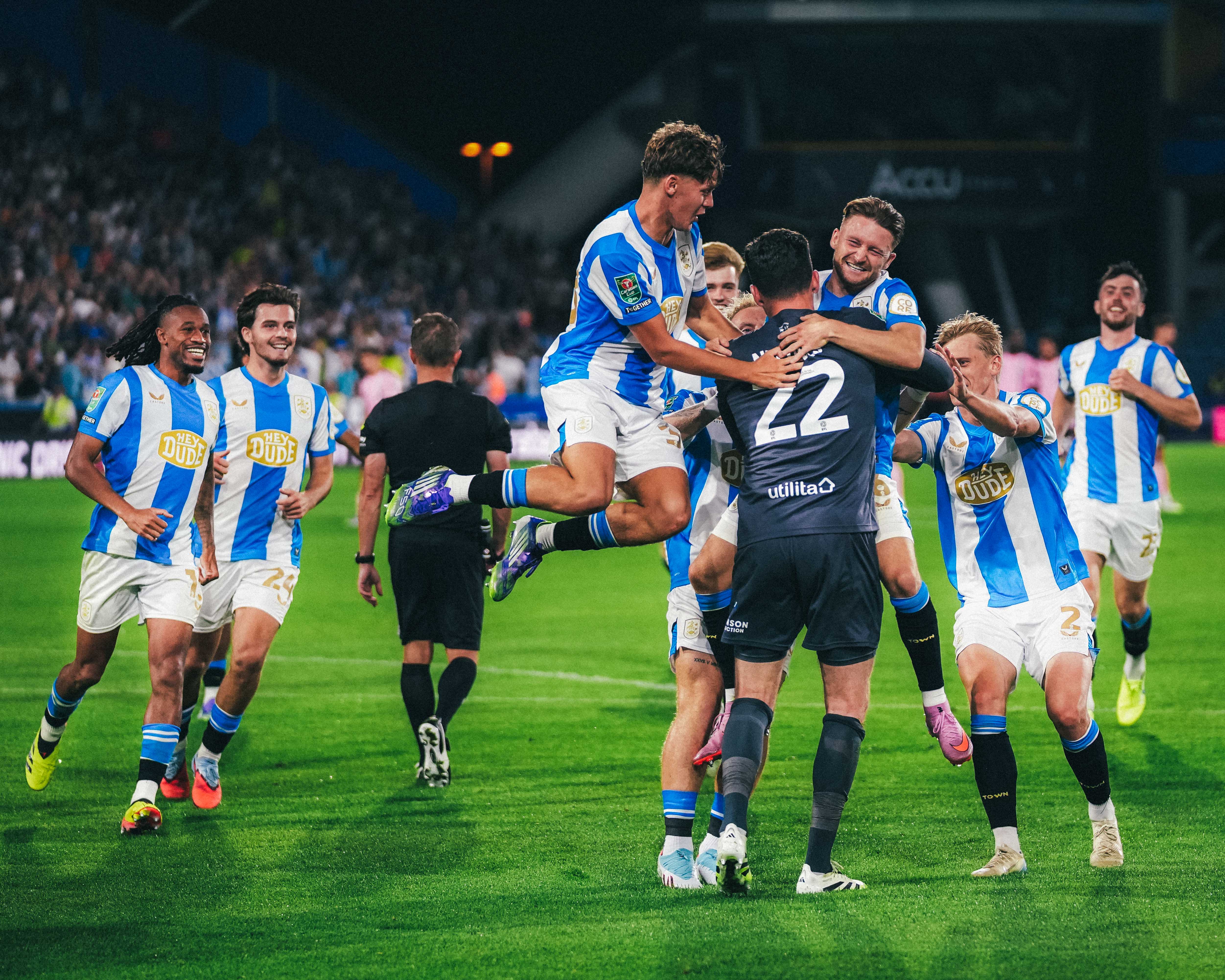 HTAFC players embrace in joy after beating LCFC on penalties and going through to the next round of the Carabao cup under floodlights. They all gather round and jump on the shootout hero, Lee Nicholls