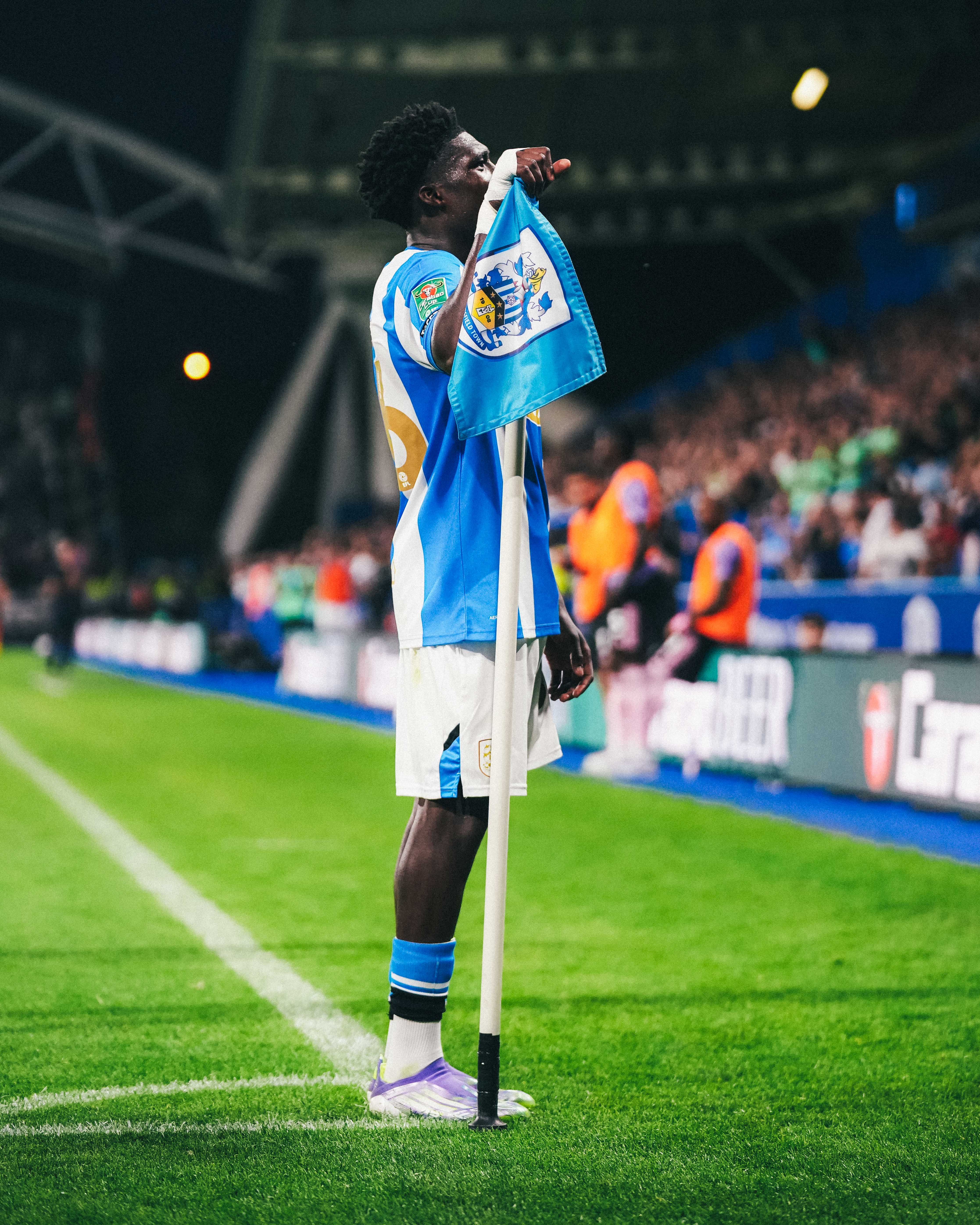 A football player in blue and white celebrates passionately at the corner flag, gripping it tightly against the backdrop of roaring fans and stadium floodlights.