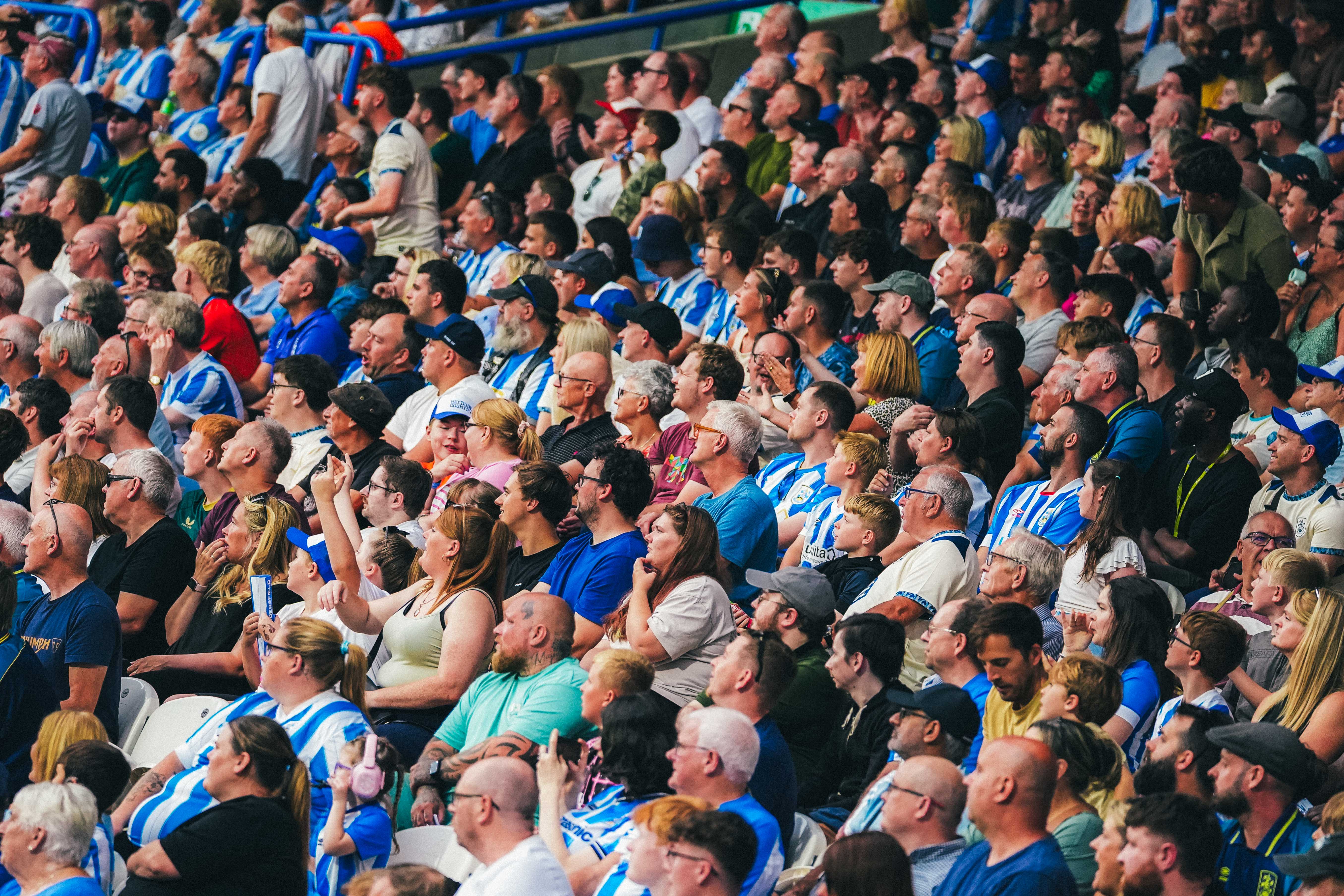 A vibrant sea of HTAFC supporters fills the stadium stands, many wearing blue and white striped jerseys, capturing the passion, anticipation, and energy of live football on match day.