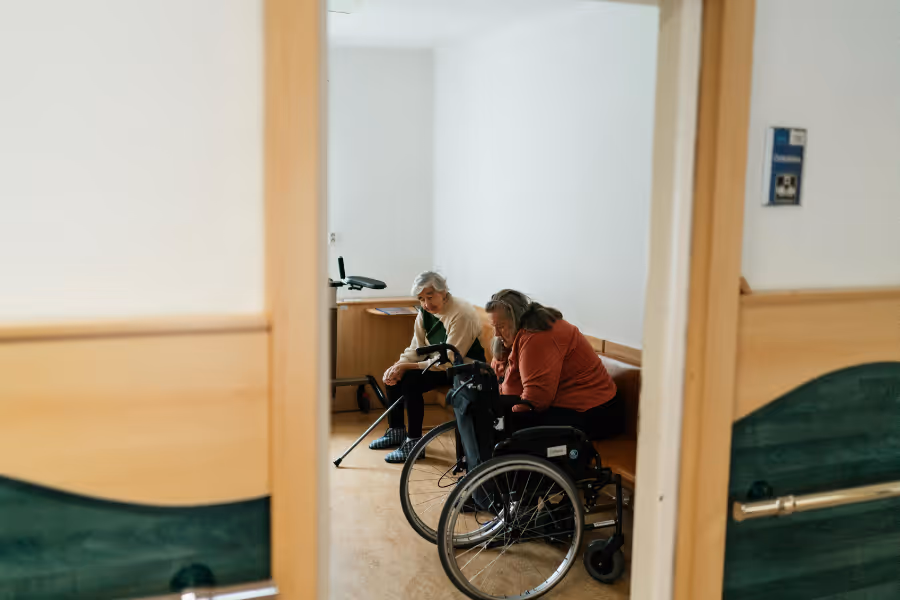 Two individuals in a waiting room, looking tired, one with a crutch, the other a wheelchair