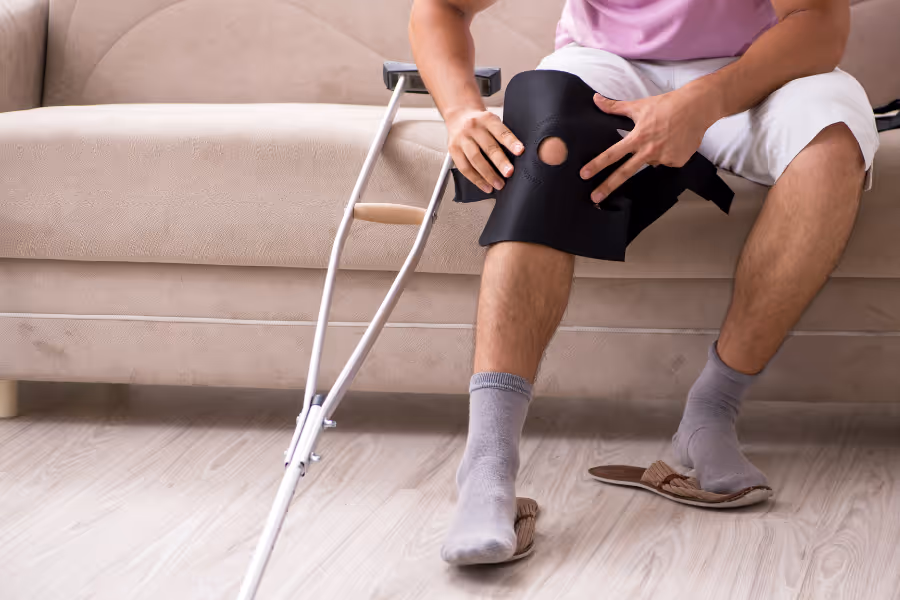 Un homme étendu sur un divan enfile une genouillère pour faciliter sa convalescence après une méniscectomie.