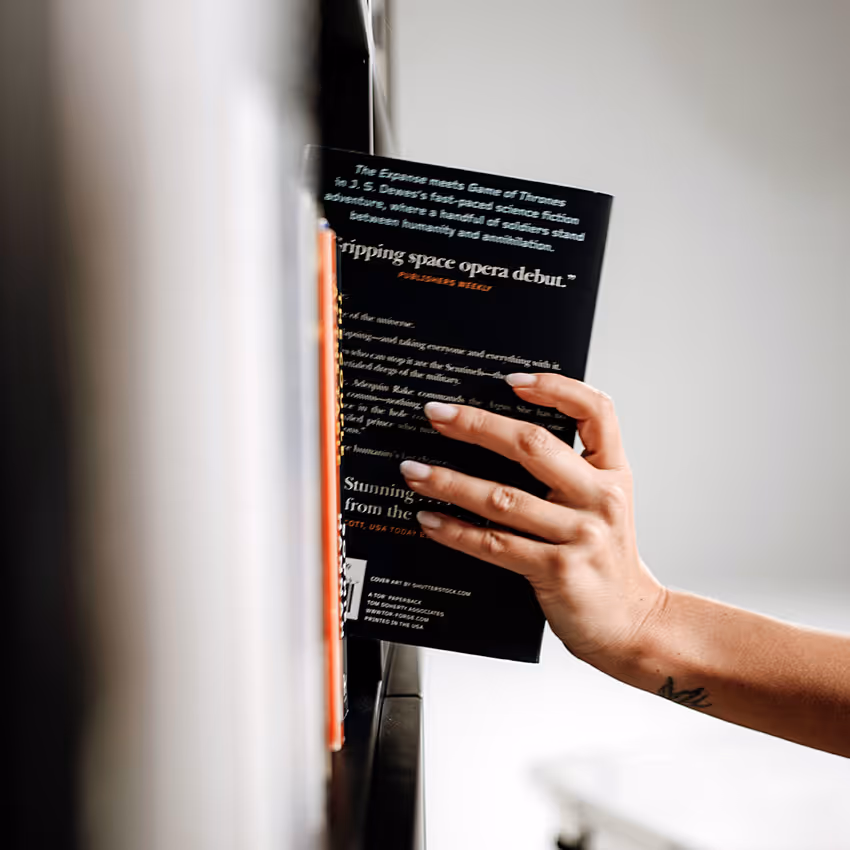 Hand with a tattoo picking a black book from a bookshelf.