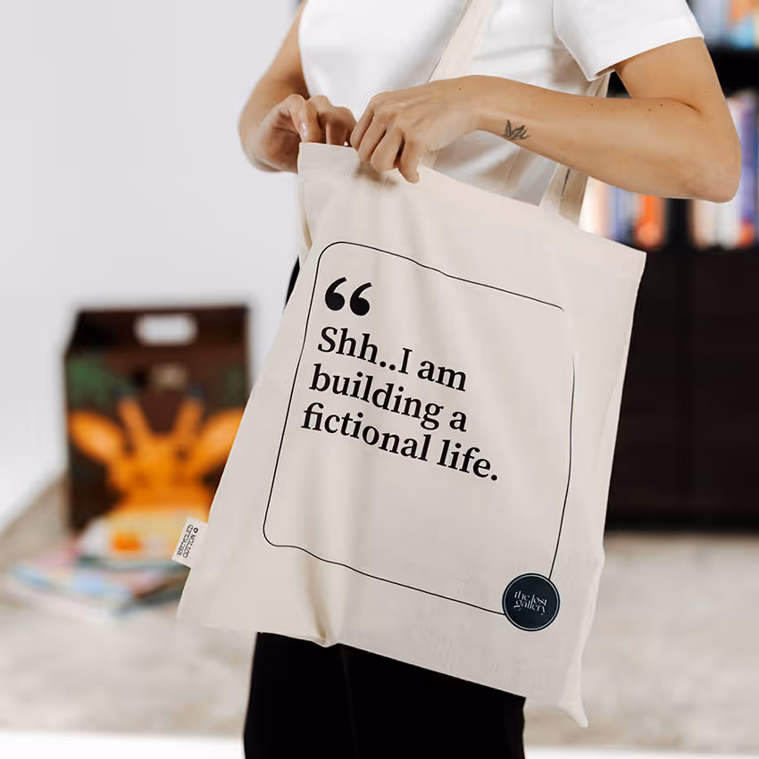 Person holding a beige tote bag with the text ‘Shh.. I am building a fictional life.’ printed on it.