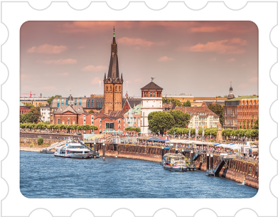 Riverfront cityscape with boats docked along the quay and a tall church spire under a cloudy sky.