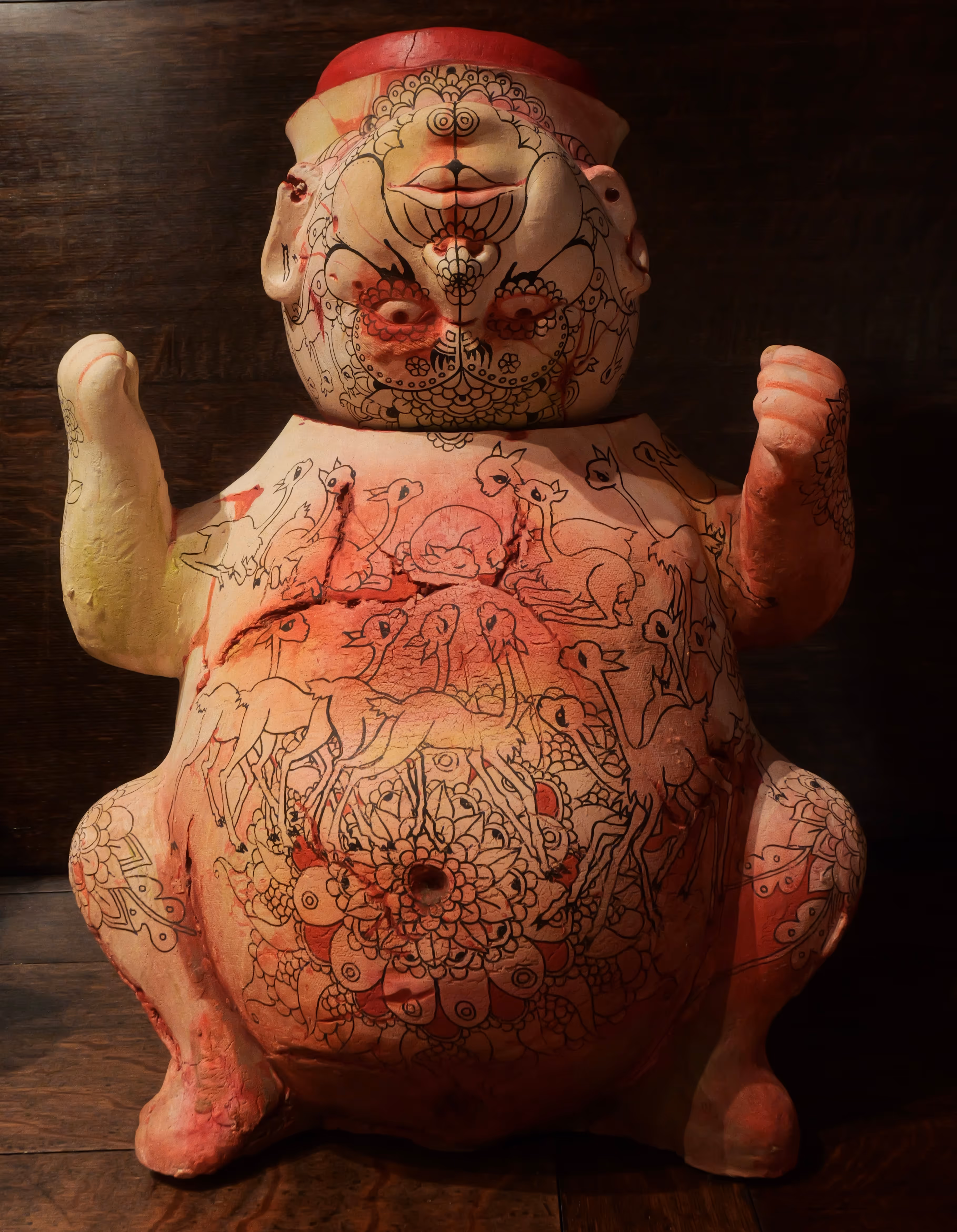 A short squat sculpt of an ornate tattooed golden figure stained orange with misshapen head and arms raised