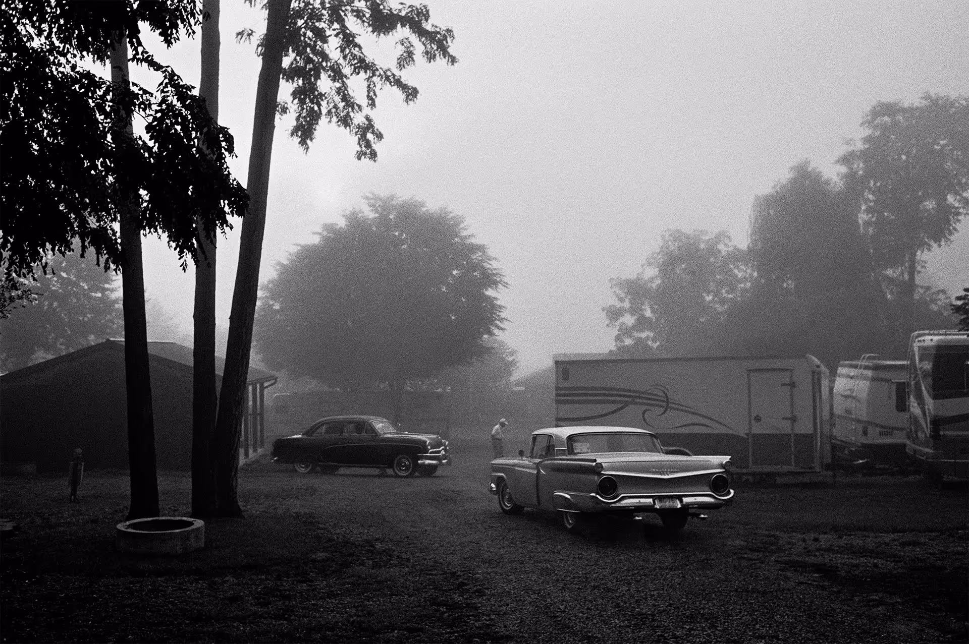 Black and white misty photo of old cars and trucks on driveway by building in tree grove