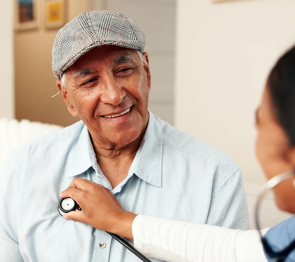 Smiling elderly man wearing a cap being examined with a stethoscope by a healthcare professional.