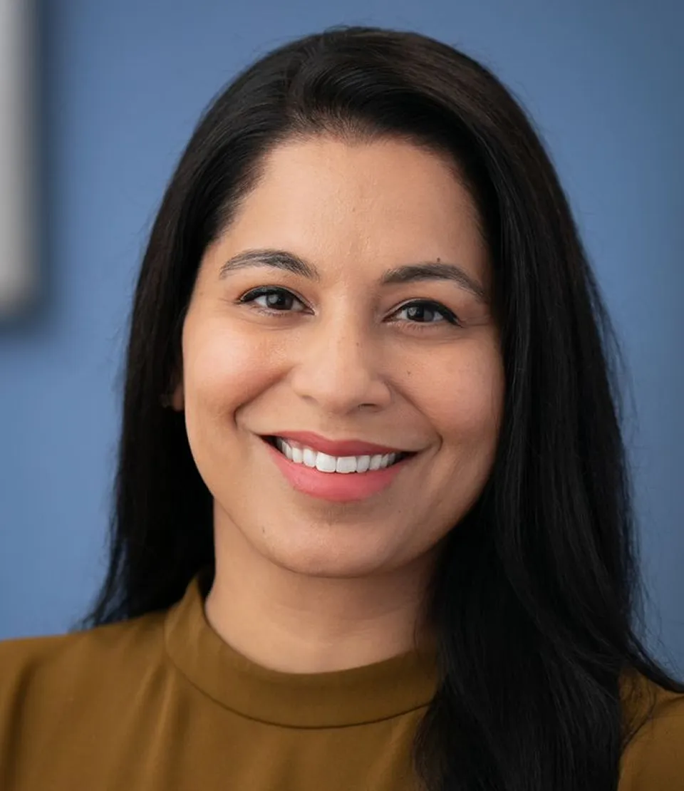 Smiling woman with long dark hair wearing a brown top against a blue background.