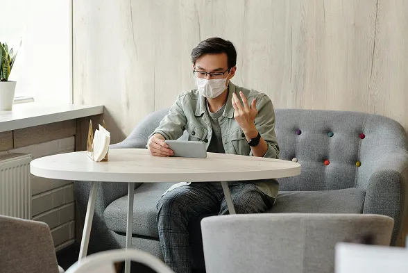 Man wearing a face mask sitting on a gray couch at a round table, holding a tablet and gesturing with his hand.