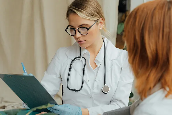 Female doctor wearing glasses and a stethoscope reviews a clipboard, consulting with a patient.