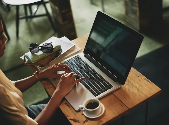 Person typing on a laptop at a wooden table with a cup of coffee, sunglasses, and books nearby.